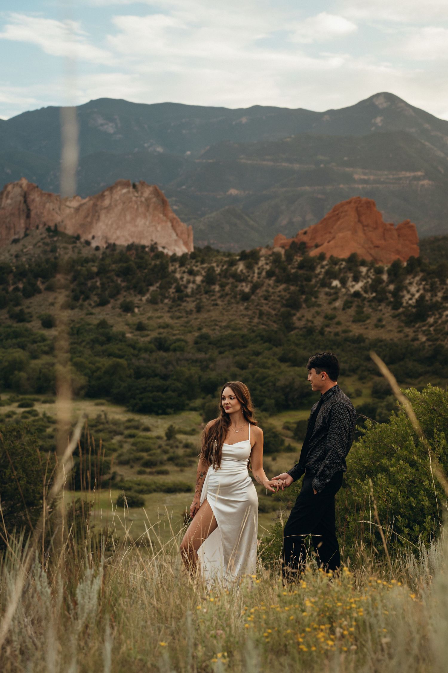A couple walks through tall grass with the majestic red rocks of Garden of the Gods and mountains in the background.