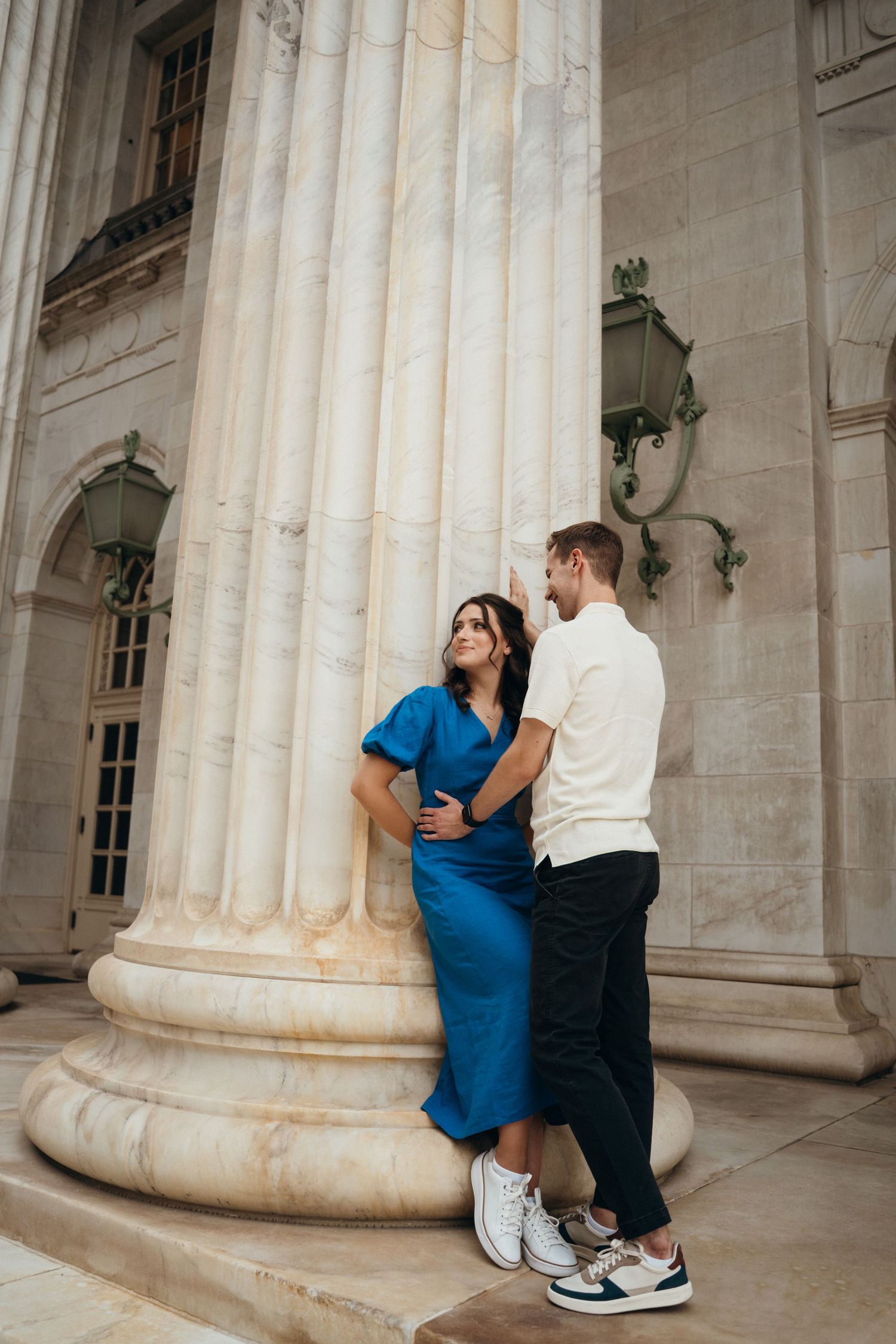 A couple in elegant attire poses by classic marble columns, with the person in a blue dress leaning against the pillar.
