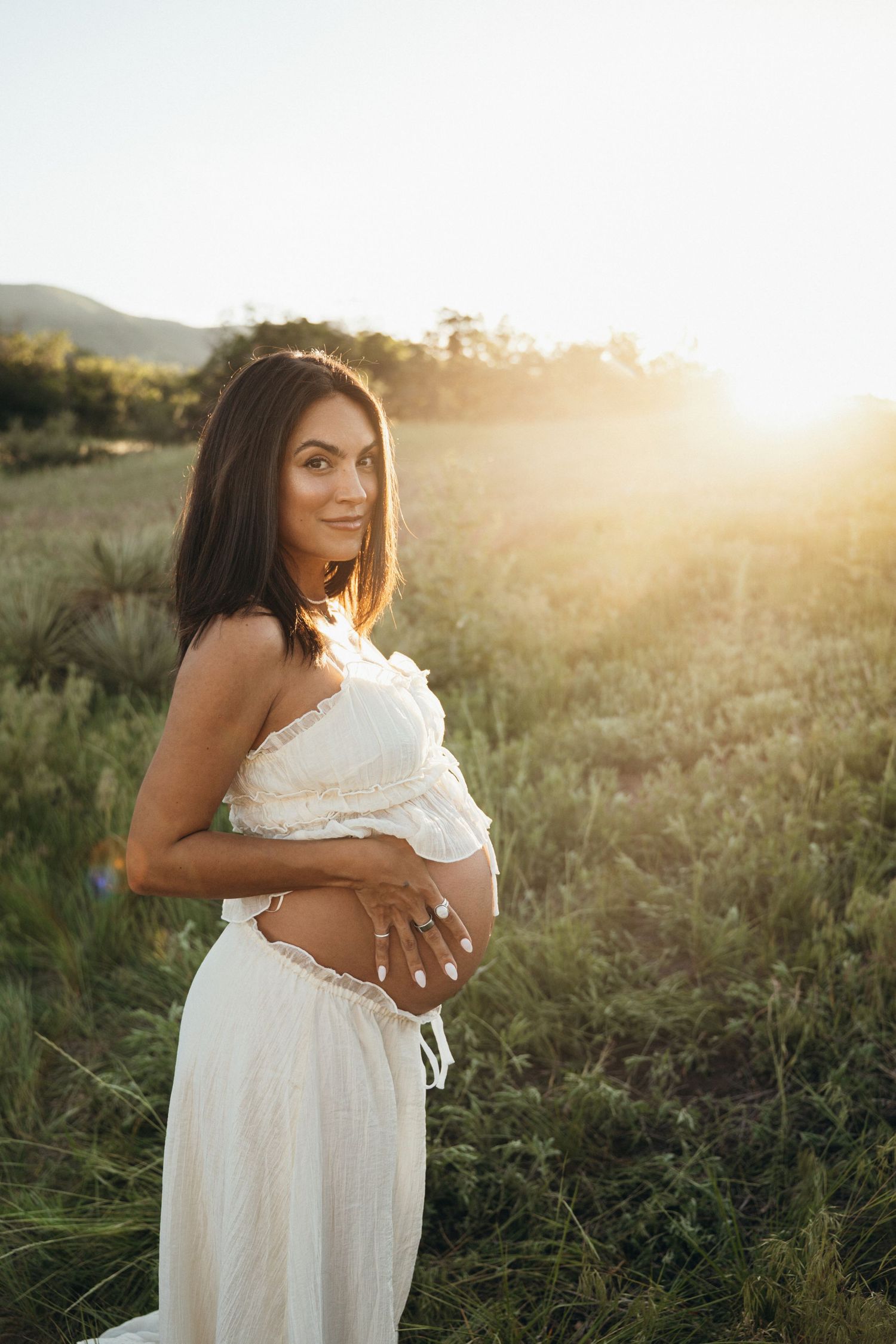 A pregnant woman in white dress poses in a field during golden hour sunset with mountains in background.
