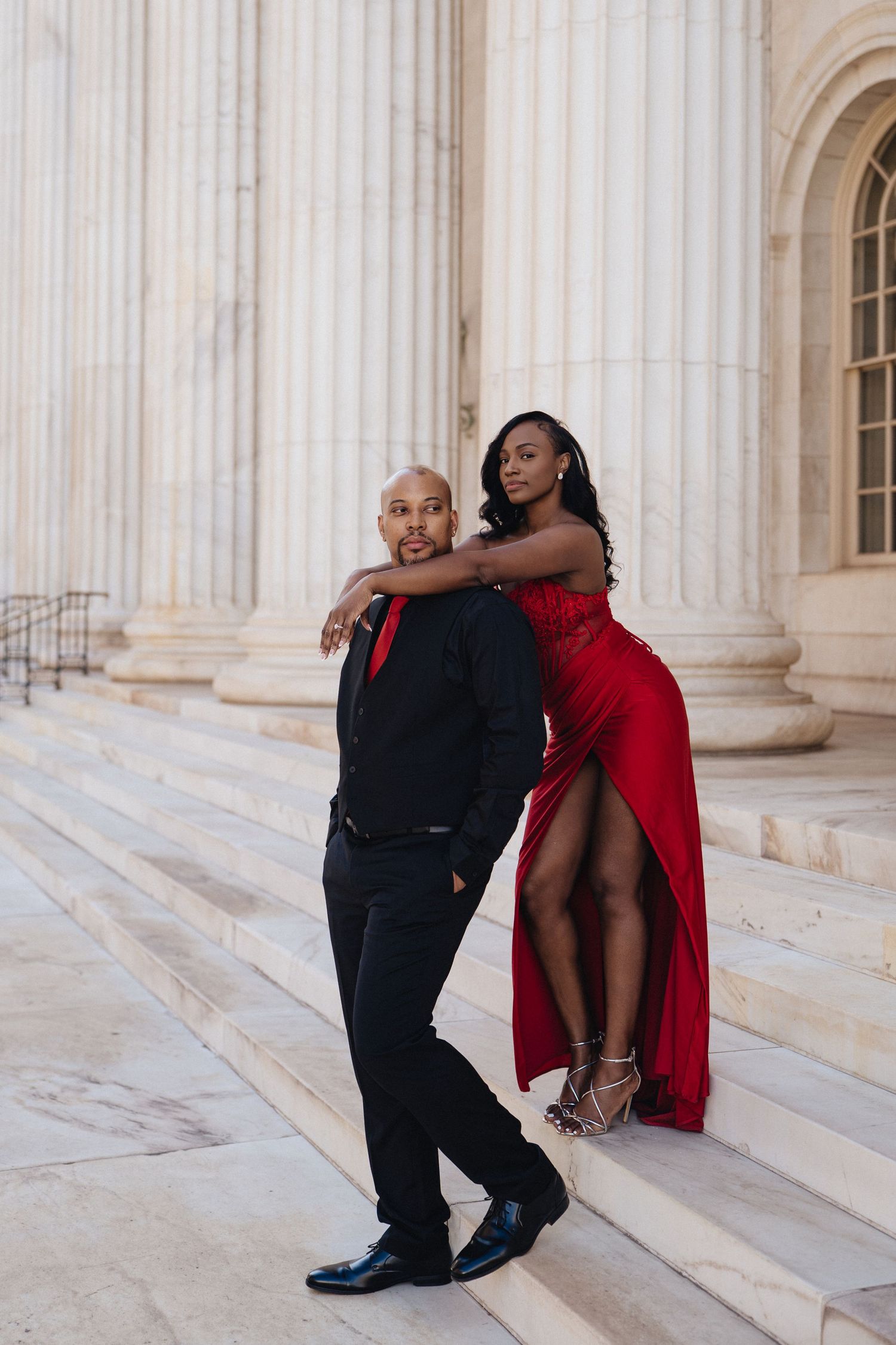Elegant couple posing on marble steps, man in black suit with red tie and woman in red slit gown.