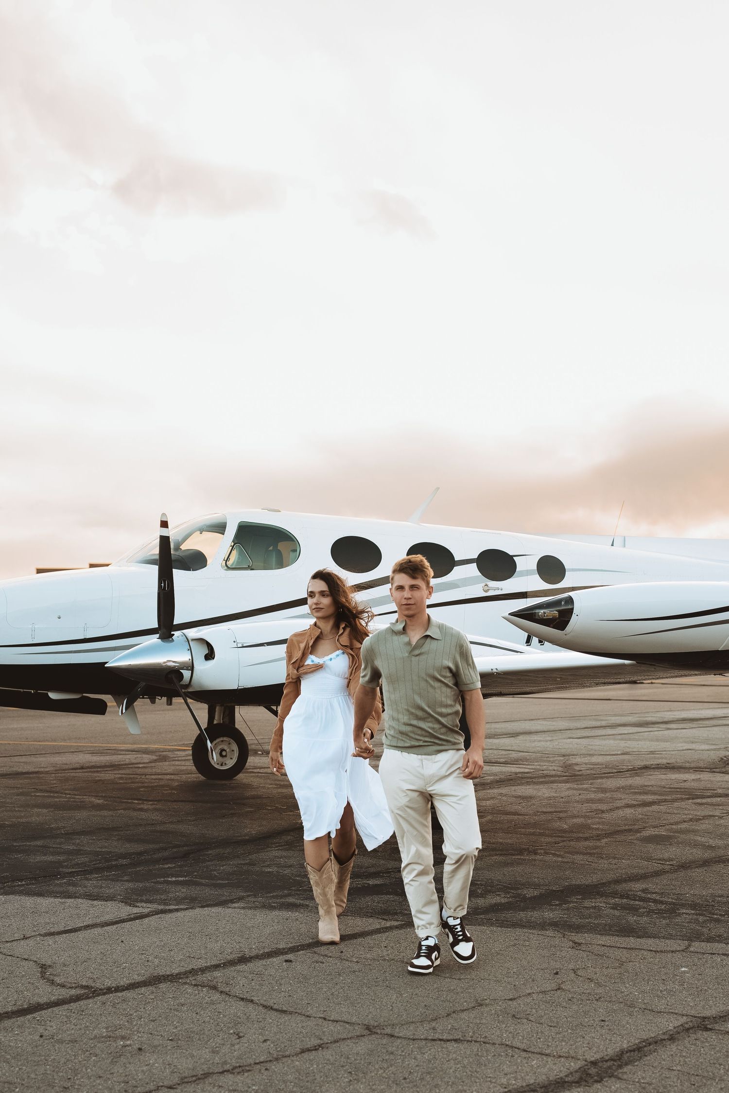 Stylishly dressed couple walking on tarmac alongside small private plane at sunset.