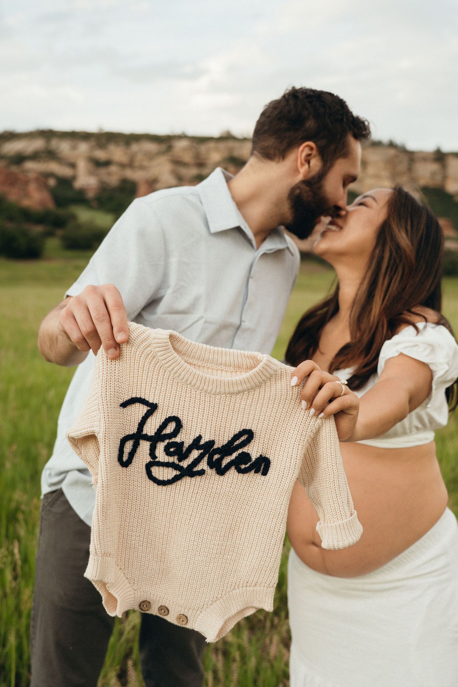 Romantic couple shares intimate moments while holding a pale beige baby onesie with embroidered text in outdoor setting.