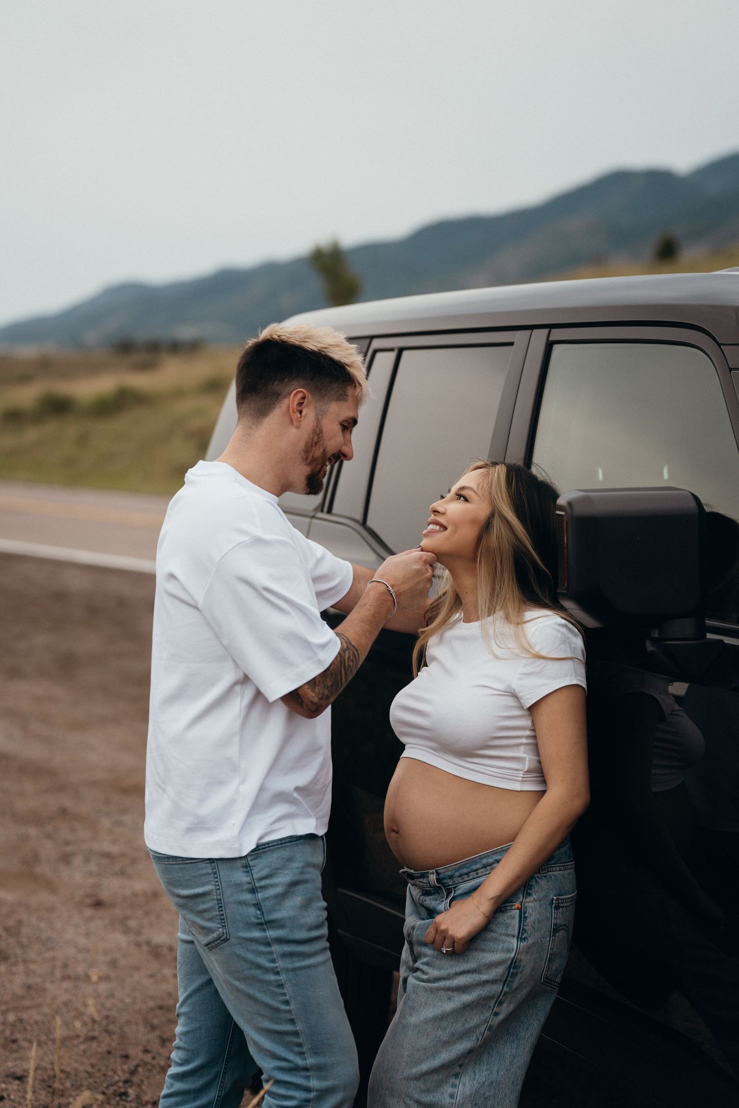 Expecting couple shares a tender moment next to their black vehicle on a rural mountain road.