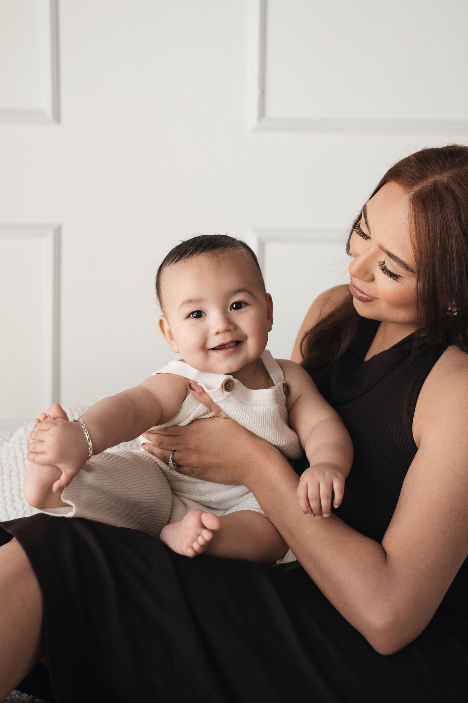 A loving caregiver holds a smiling baby wearing white while sitting against a white paneled wall.