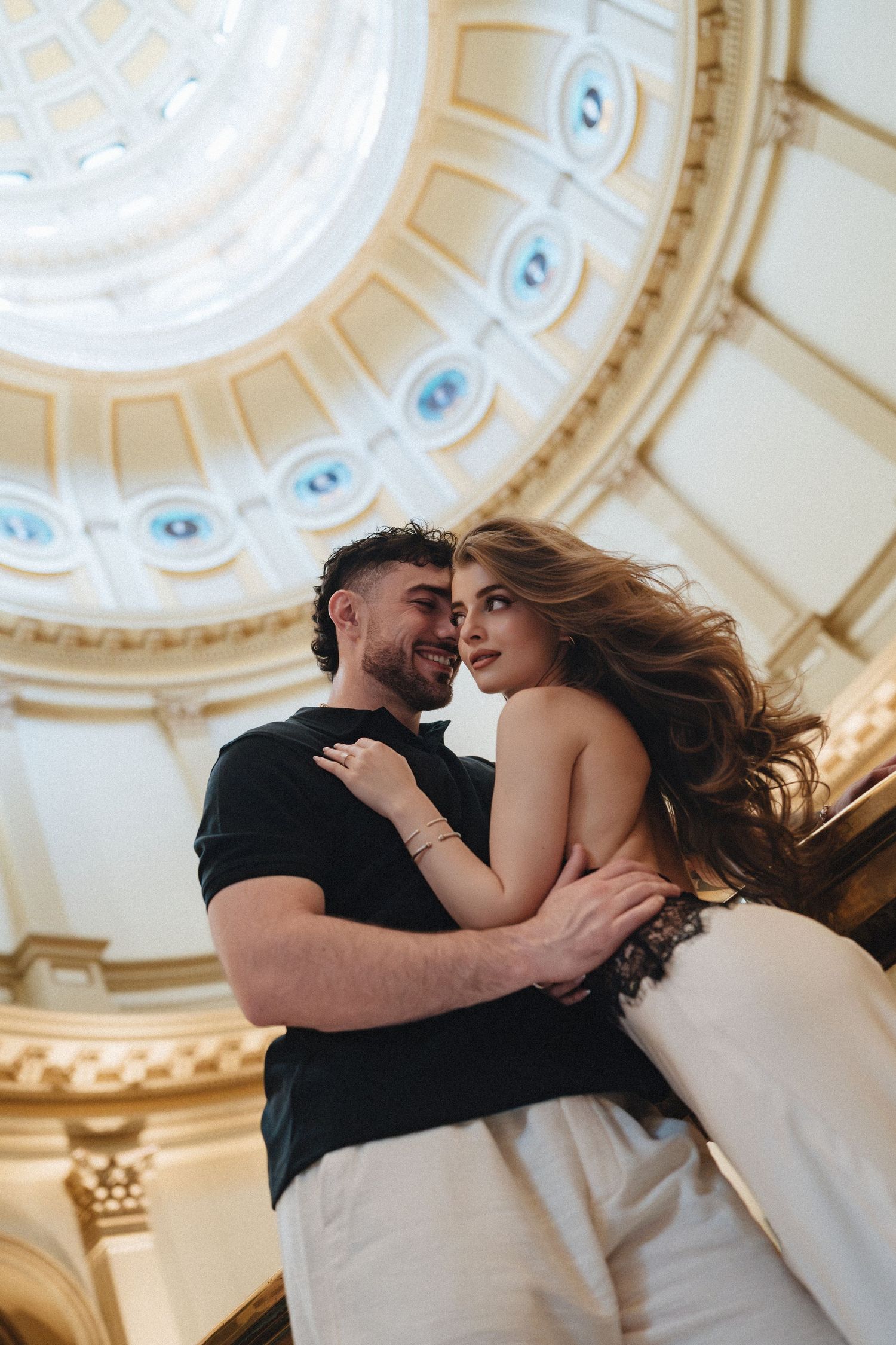 Romantic couple embracing under an ornate domed ceiling with classical architecture and decorative blue medallions.