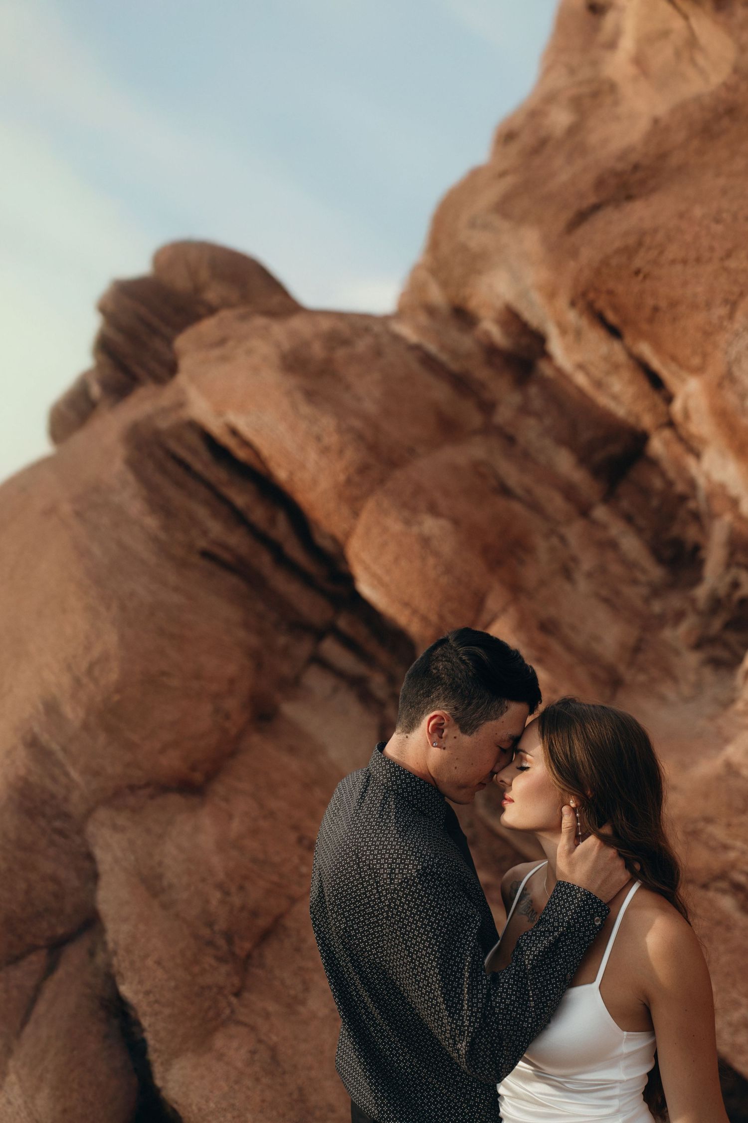Two people embrace intimately against towering red rock formations under a blue desert sky.