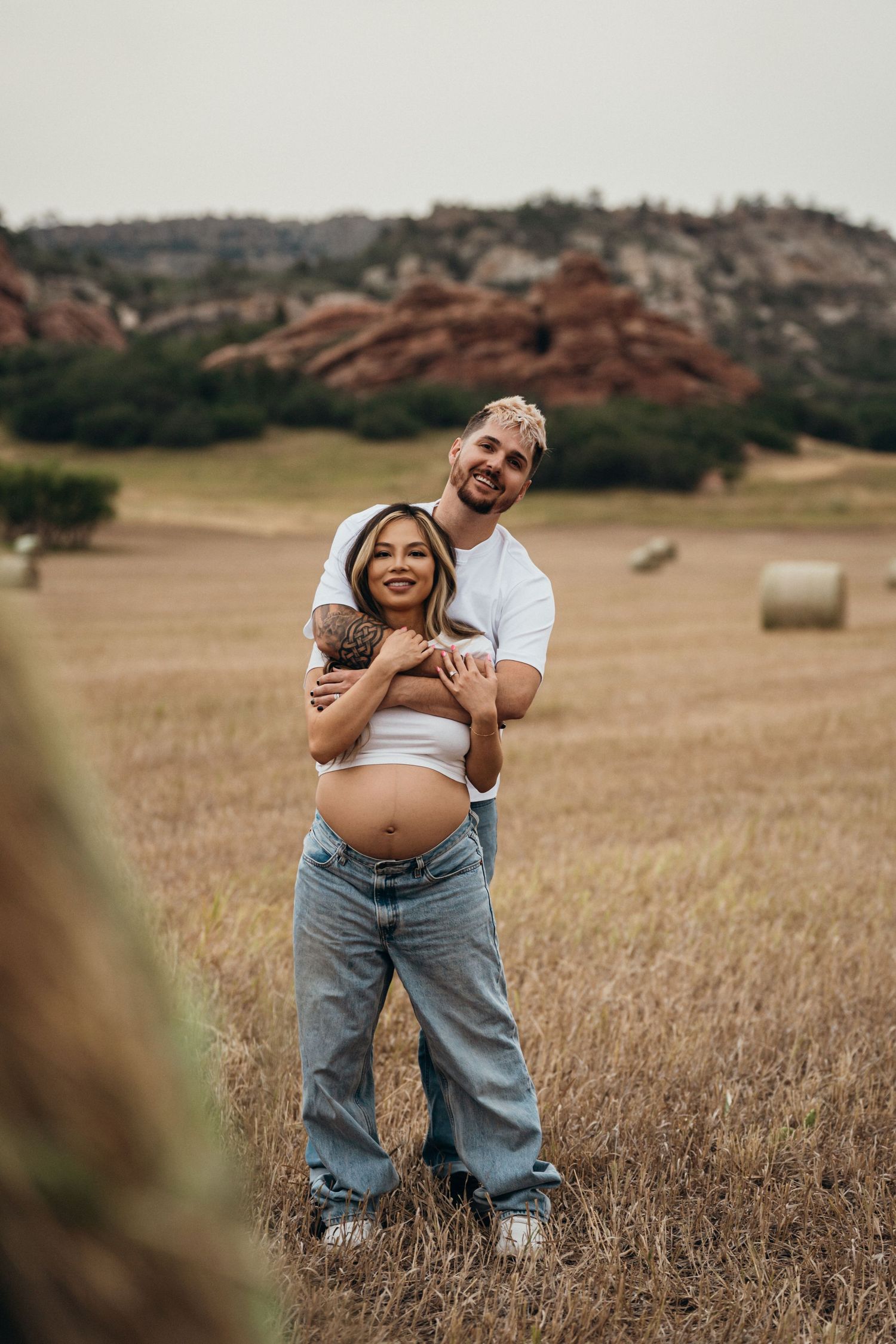 A couple embraces in a rustic field with red rock formations in the background during their maternity session.