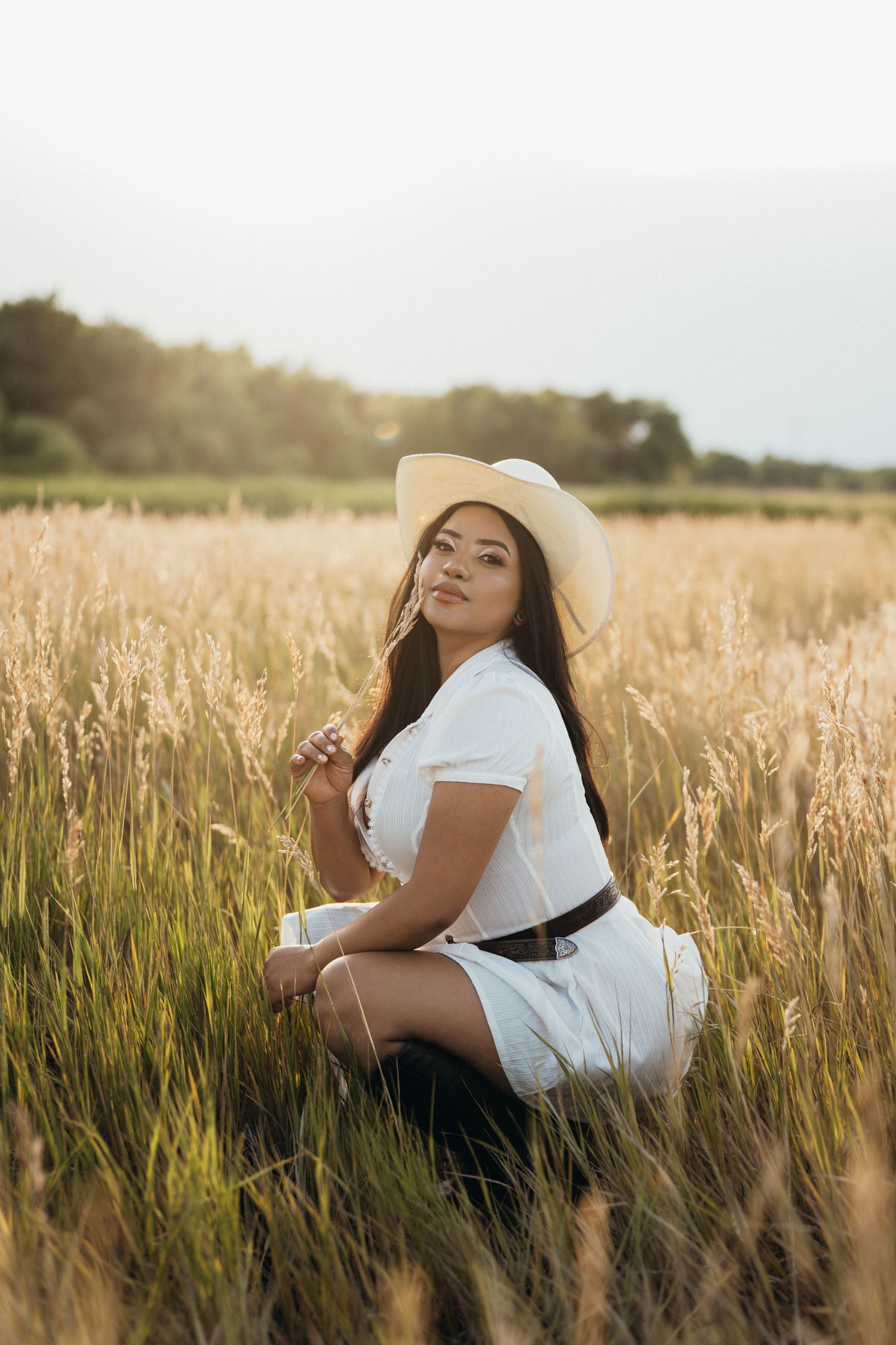 A woman in a white dress and sun hat poses in a golden wheat field during sunset.