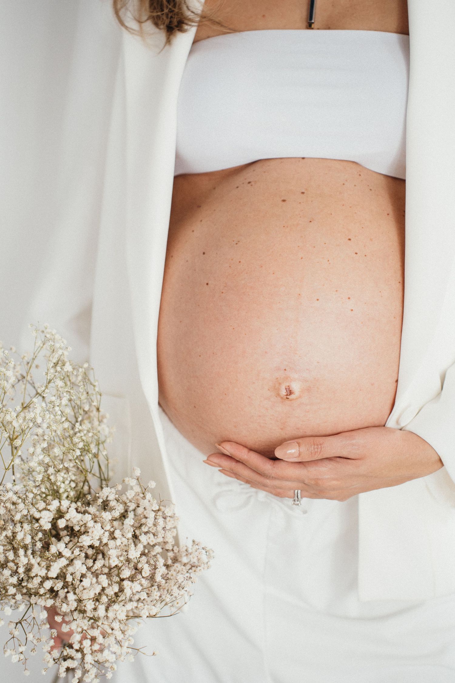 Close up of pregnant belly in white bra and robe with baby's breath flowers, showcasing a tender maternity moment.