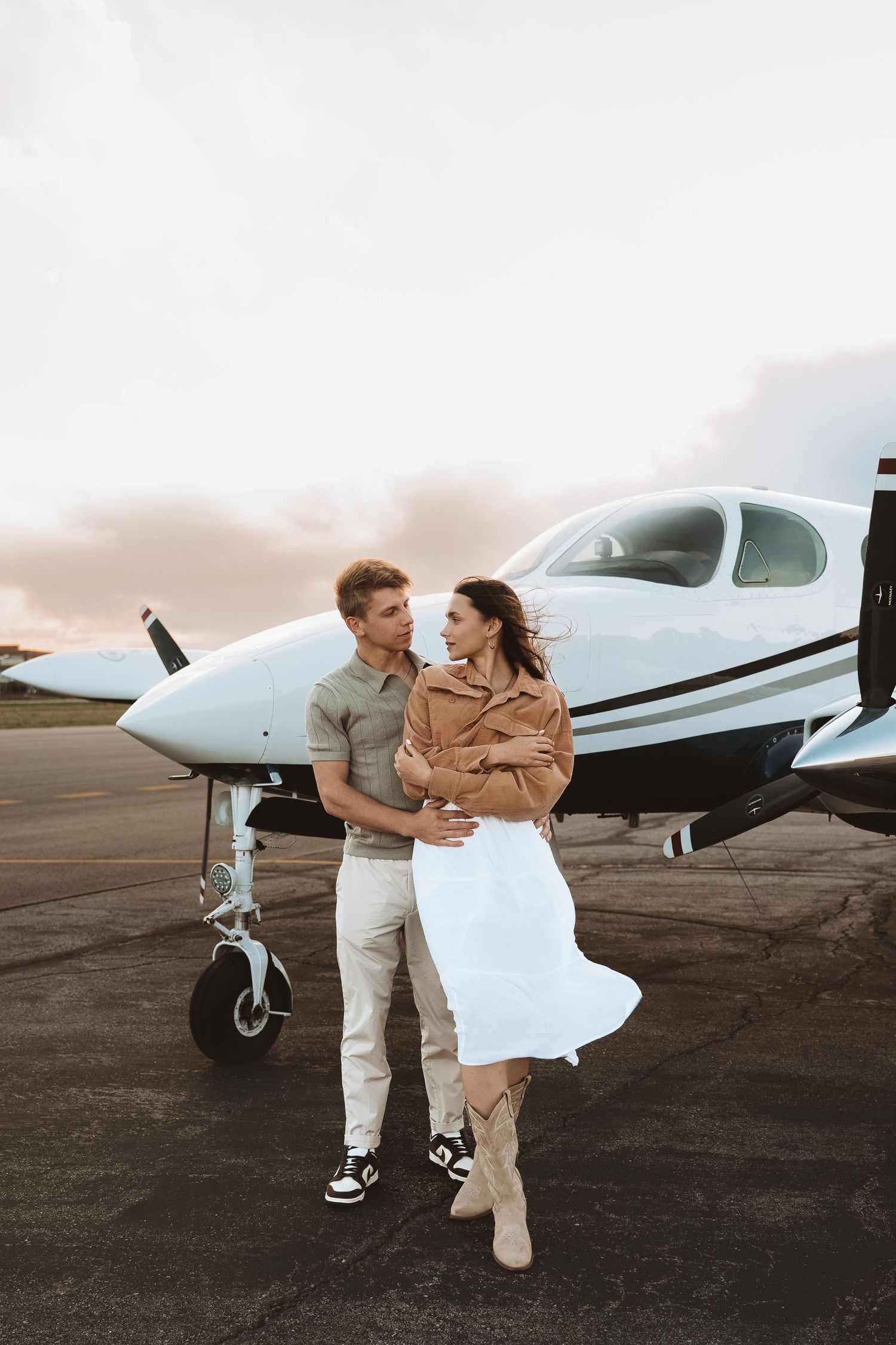 Couple stands together by a small private aircraft during a romantic photo shoot at an airport.