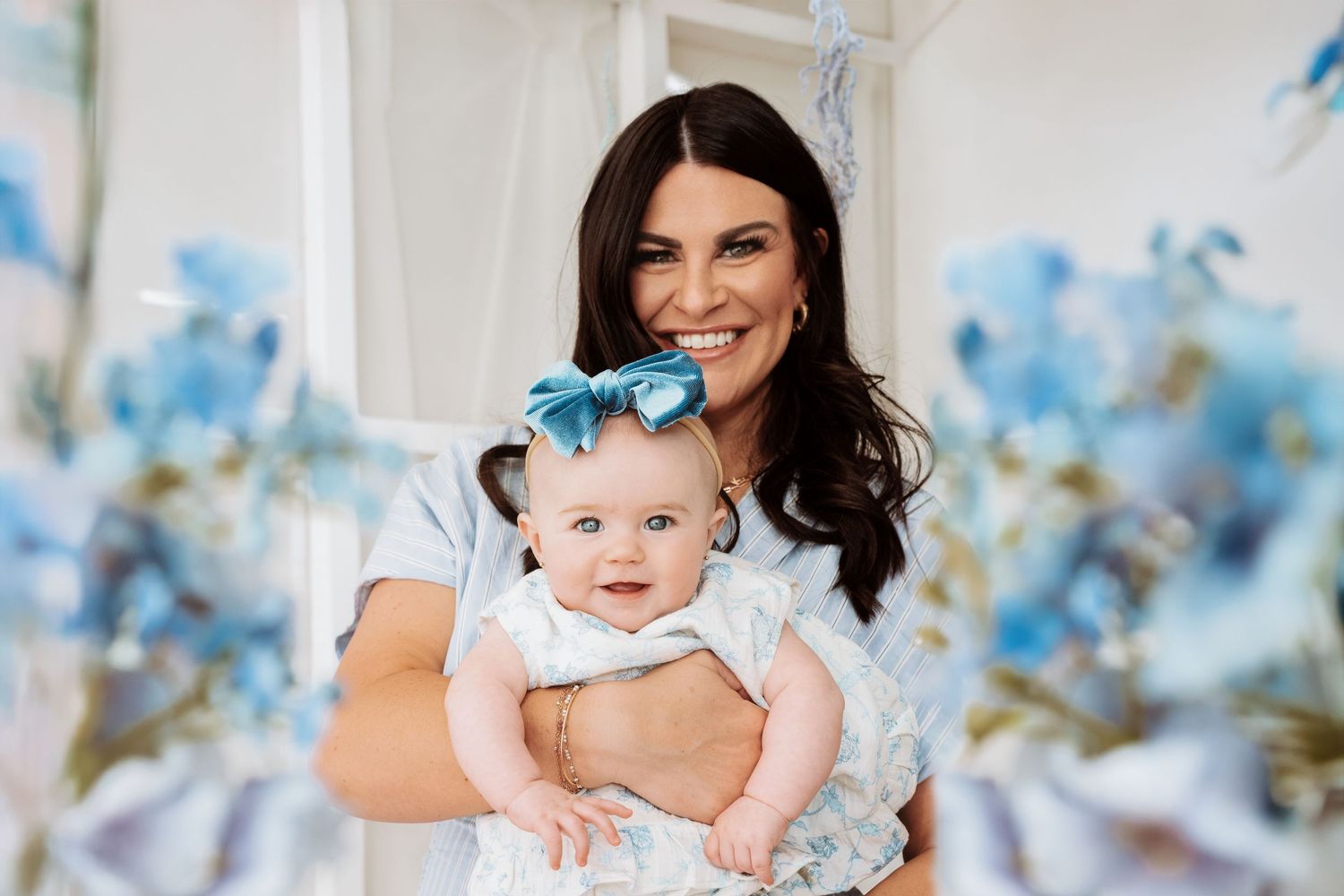 Portrait with blue floral decorations features an adult holding a baby wearing a light-colored outfit in a bright setting.