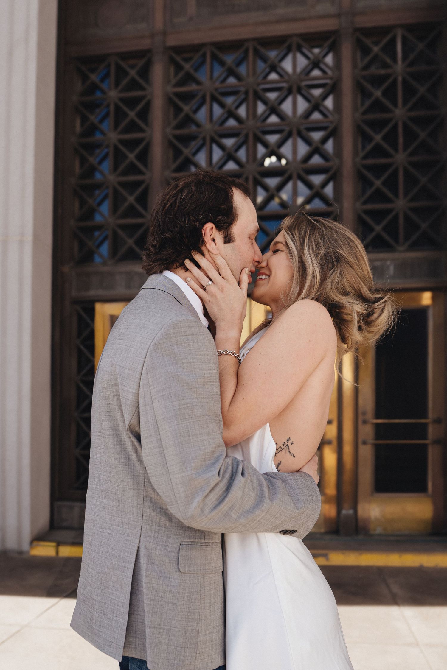 A couple in wedding attire share a romantic kiss in front of an elegant building with ornate windows.