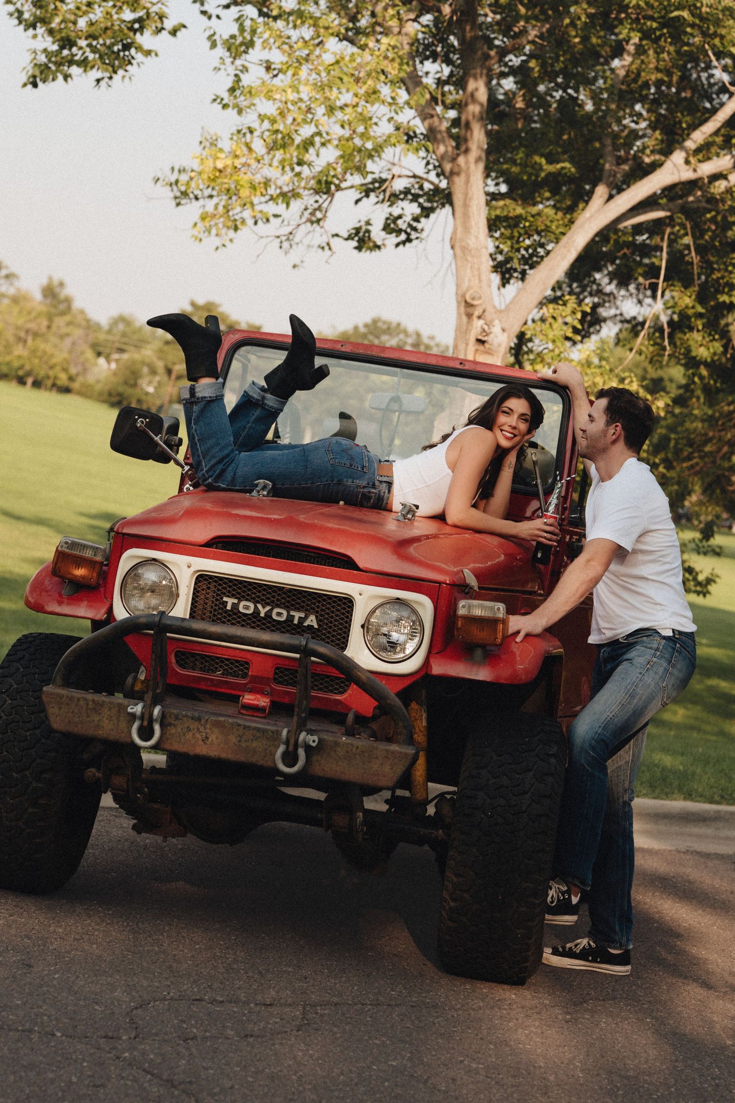 Couple posing with a vintage red Toyota FJ40 jeep outdoors, woman lying on hood, man leaning against bumper smiling.