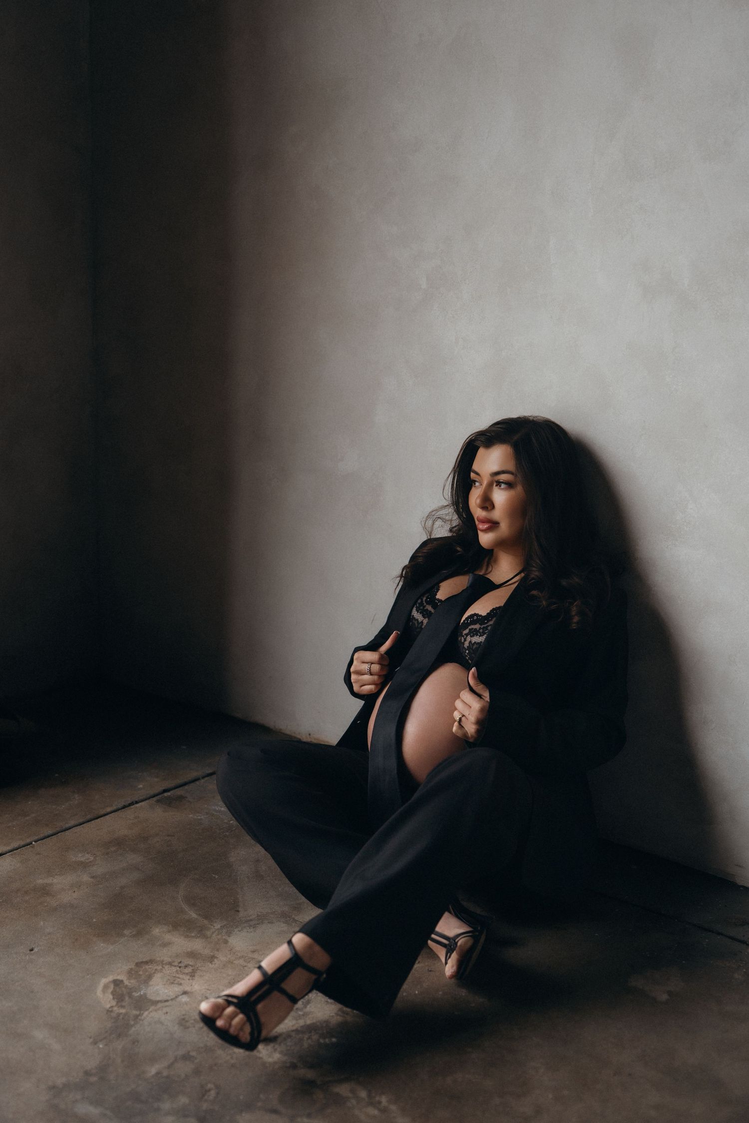 Artistic maternity portrait of woman in black outfit sitting against concrete wall in moody lighting.