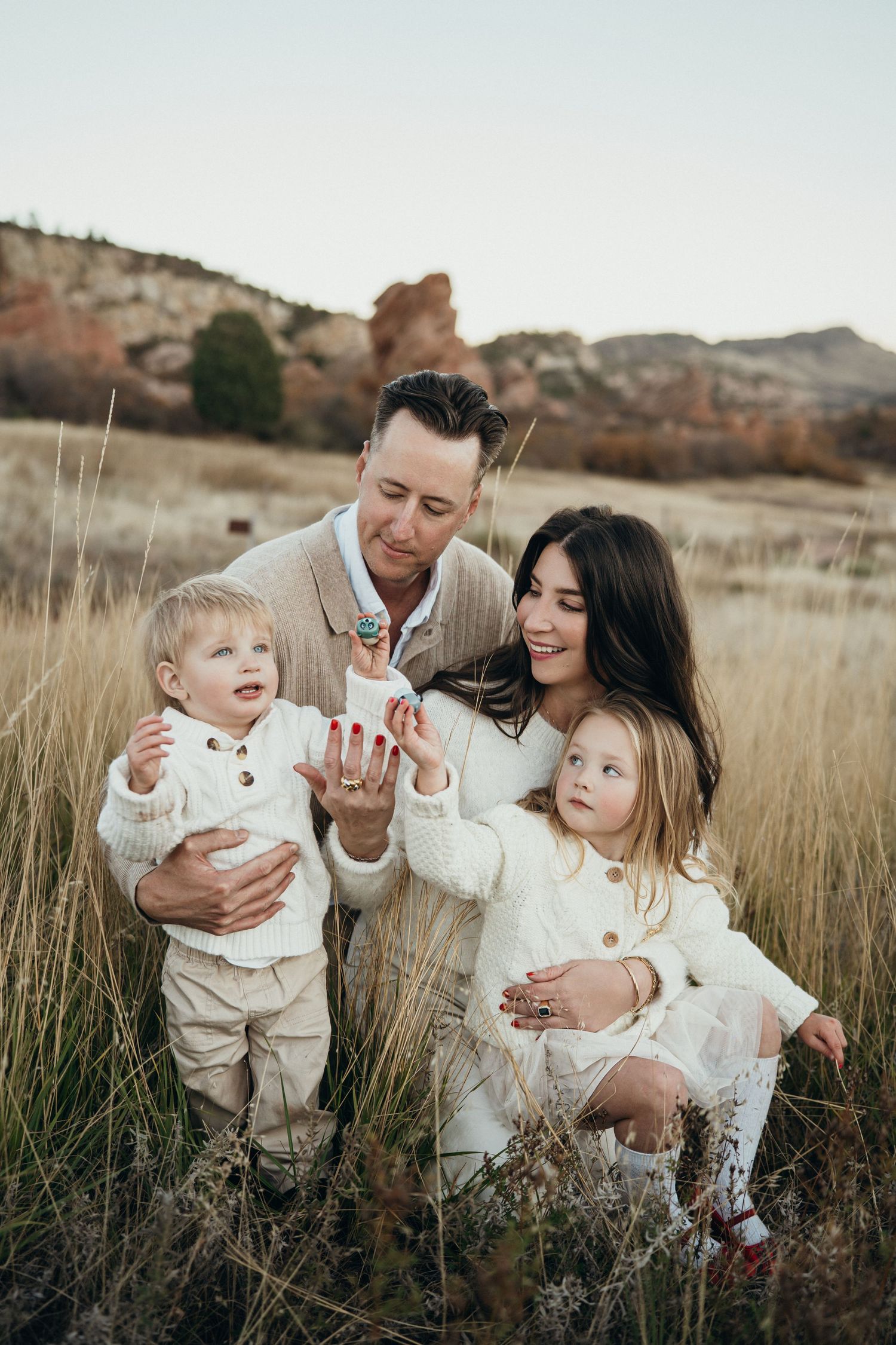 A family in cream-colored outfits poses together in a field of tall grass with mountains in the background at sunset.