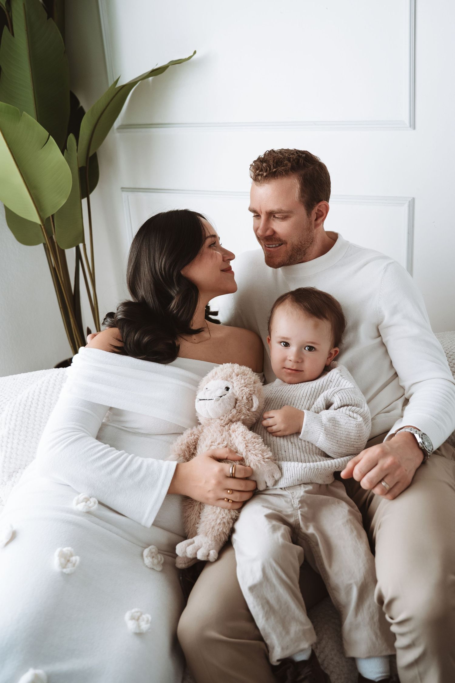 A family lounging together in white sweaters on a beige couch with a plush toy and indoor plant in background.