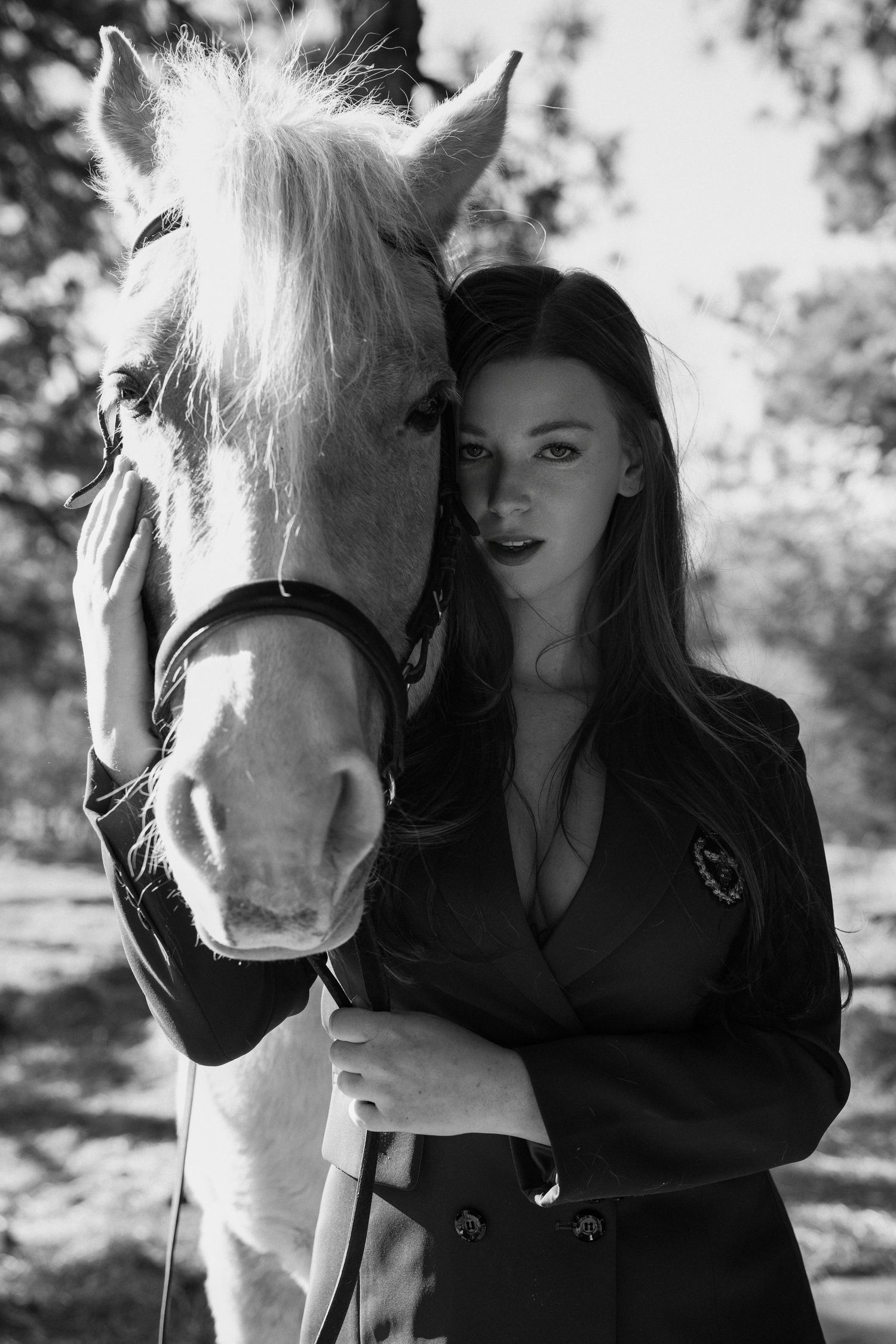 Black and white photo of a beautiful white horse and a woman in a dark blazer posing together outdoors.