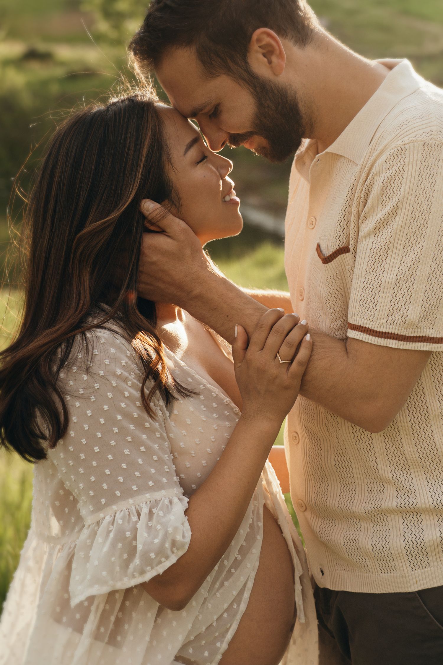 A romantic couple shares an intimate moment at sunset in a field, she in a polka dot dress and he in a cream sweater.