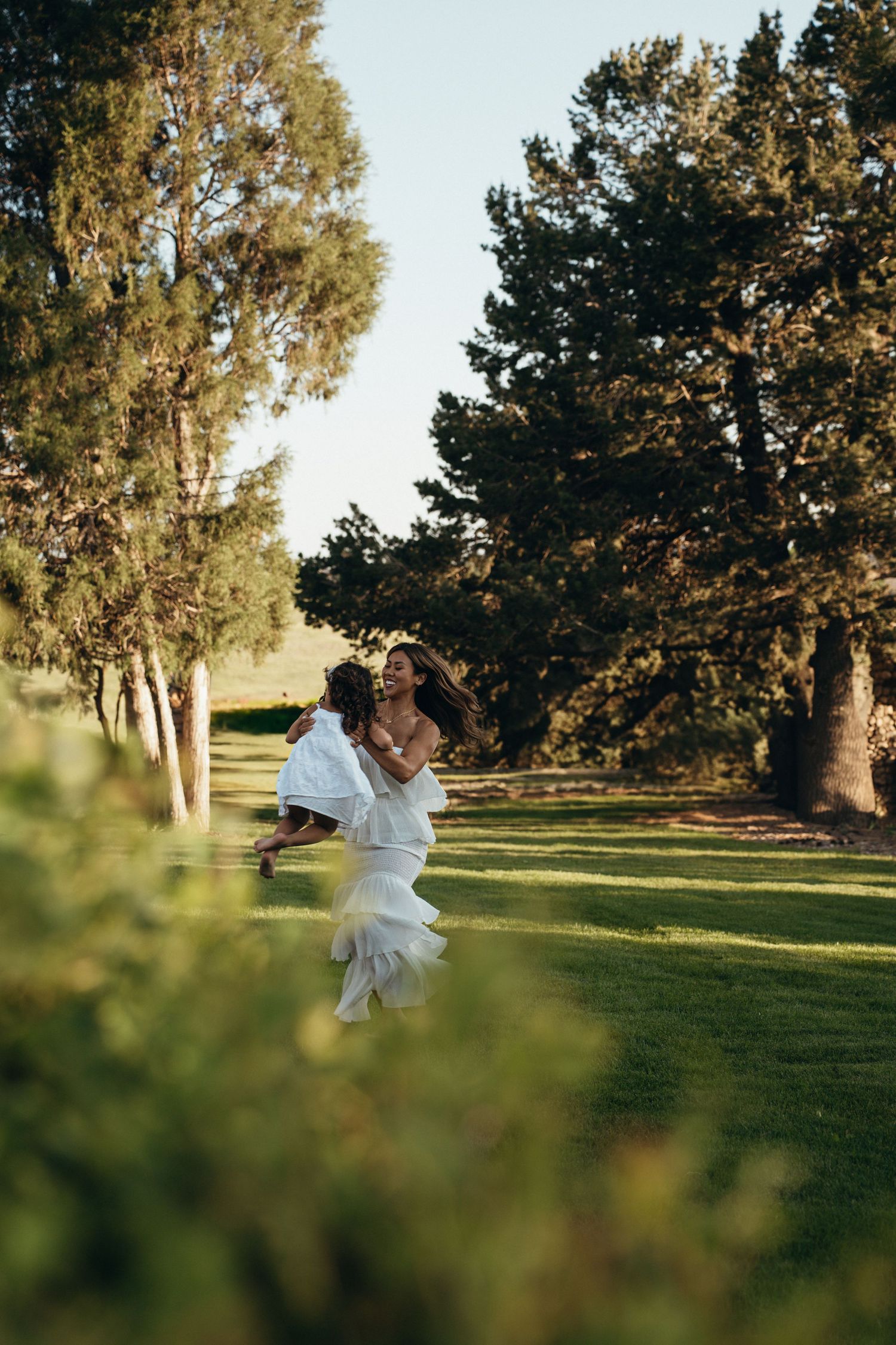 A dancer in a flowing white dress twirls gracefully between tall trees on a sunlit lawn.