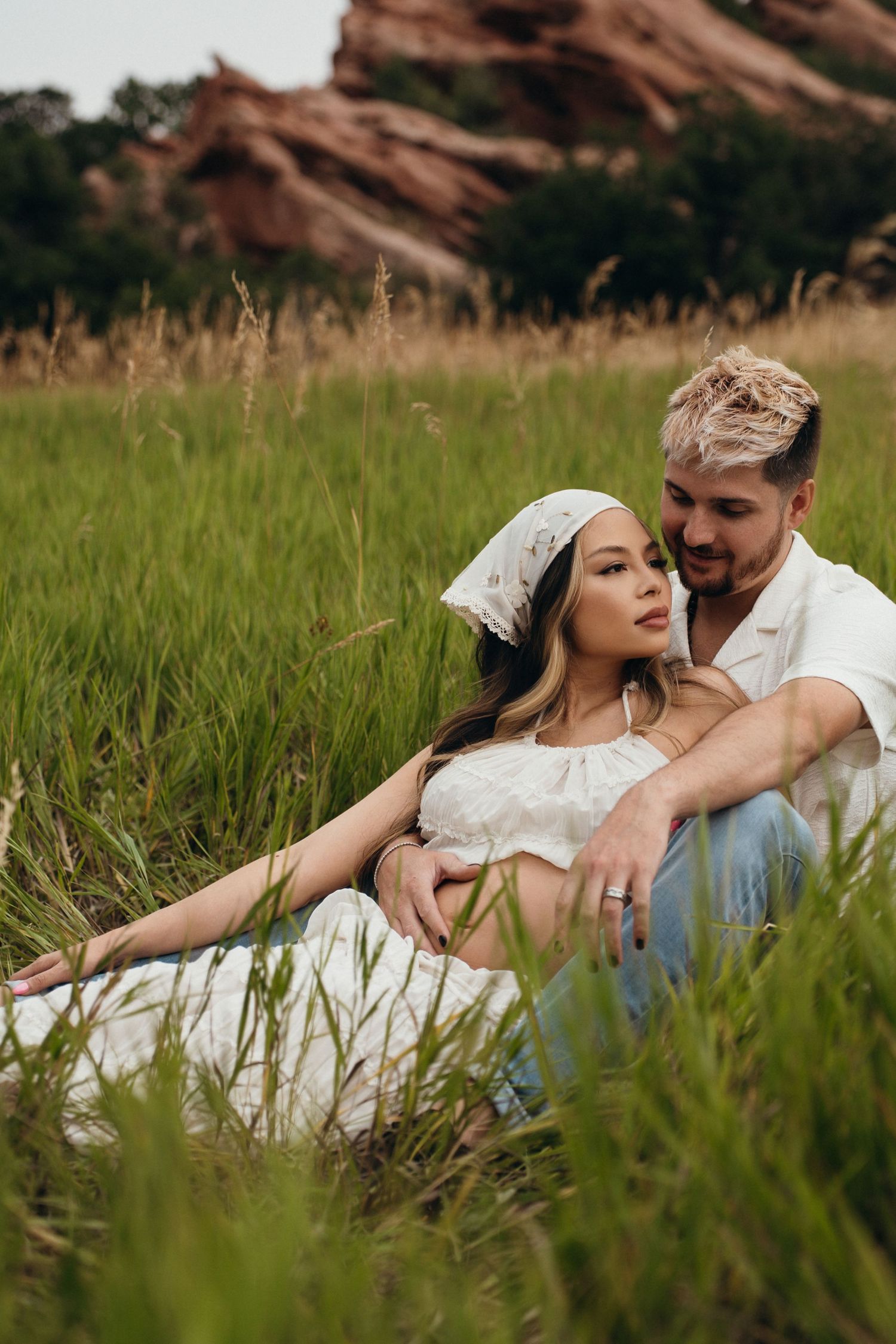 A romantic couple lounges in tall grass with red rocks and mountains visible in the background at sunset.