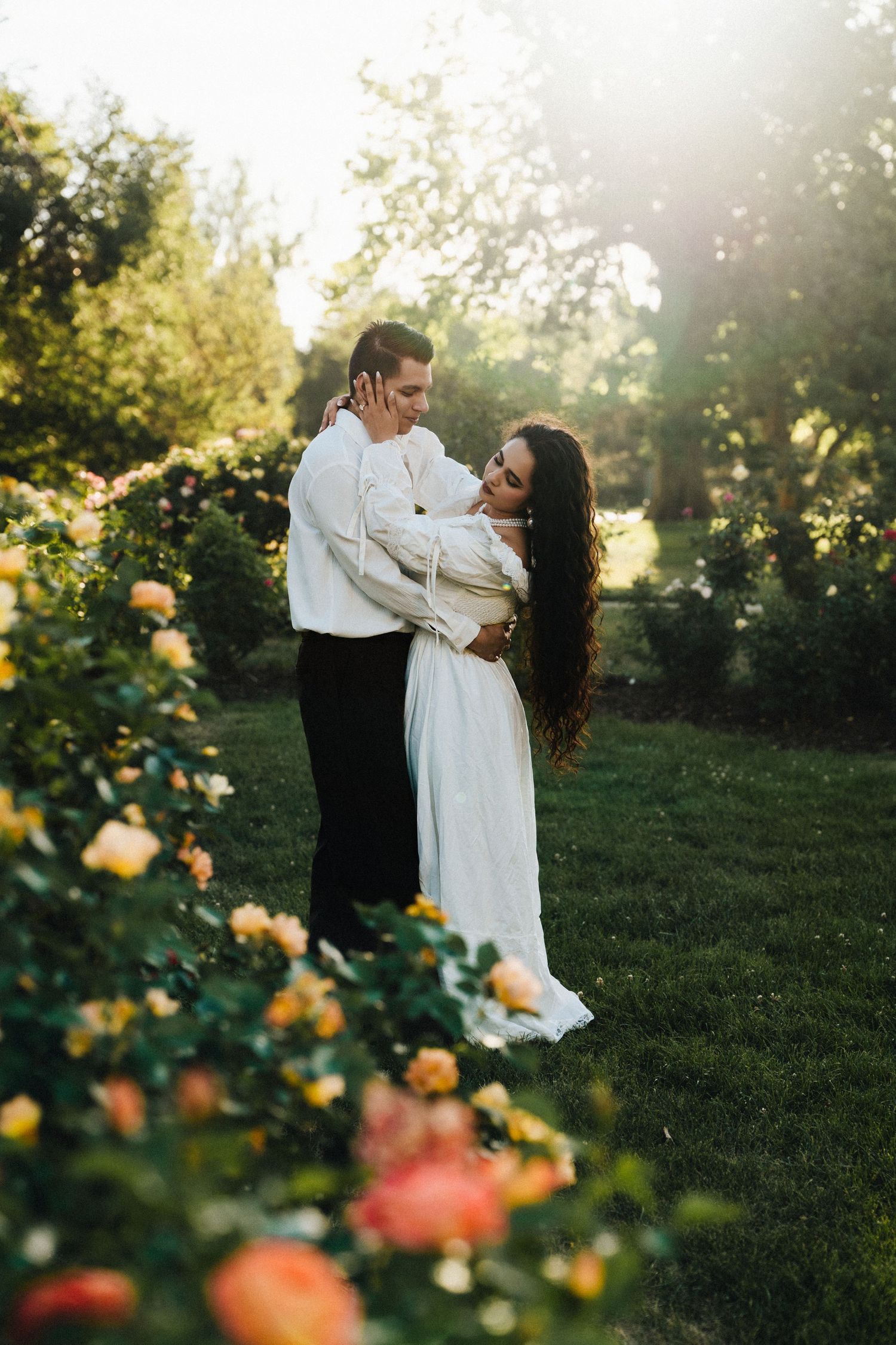 Couple embracing in a sunlit garden surrounded by blooming roses, wearing elegant white outfits.