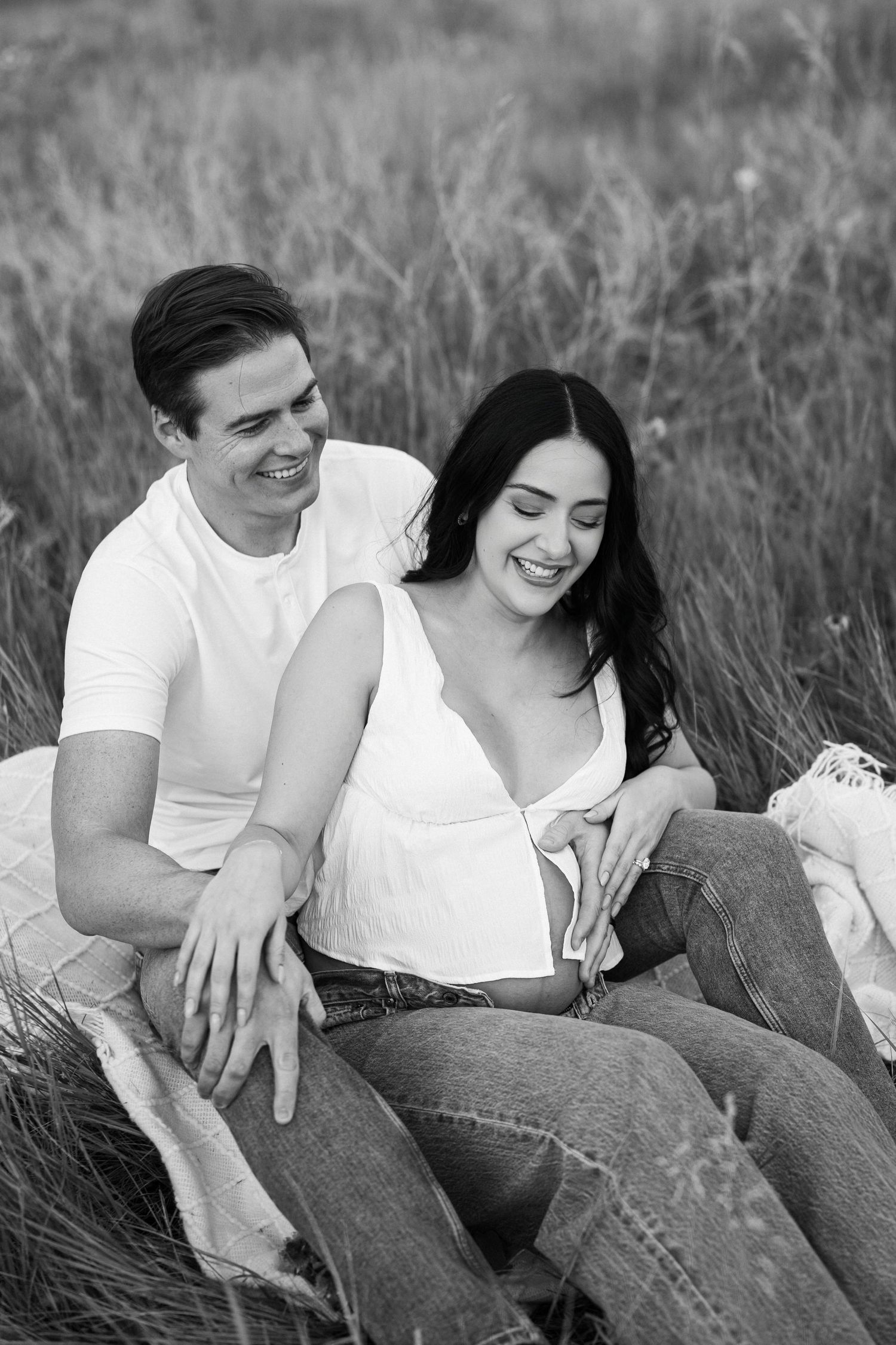 Black and white photo series of a couple sitting together in a grassy field wearing casual white tops and jeans.