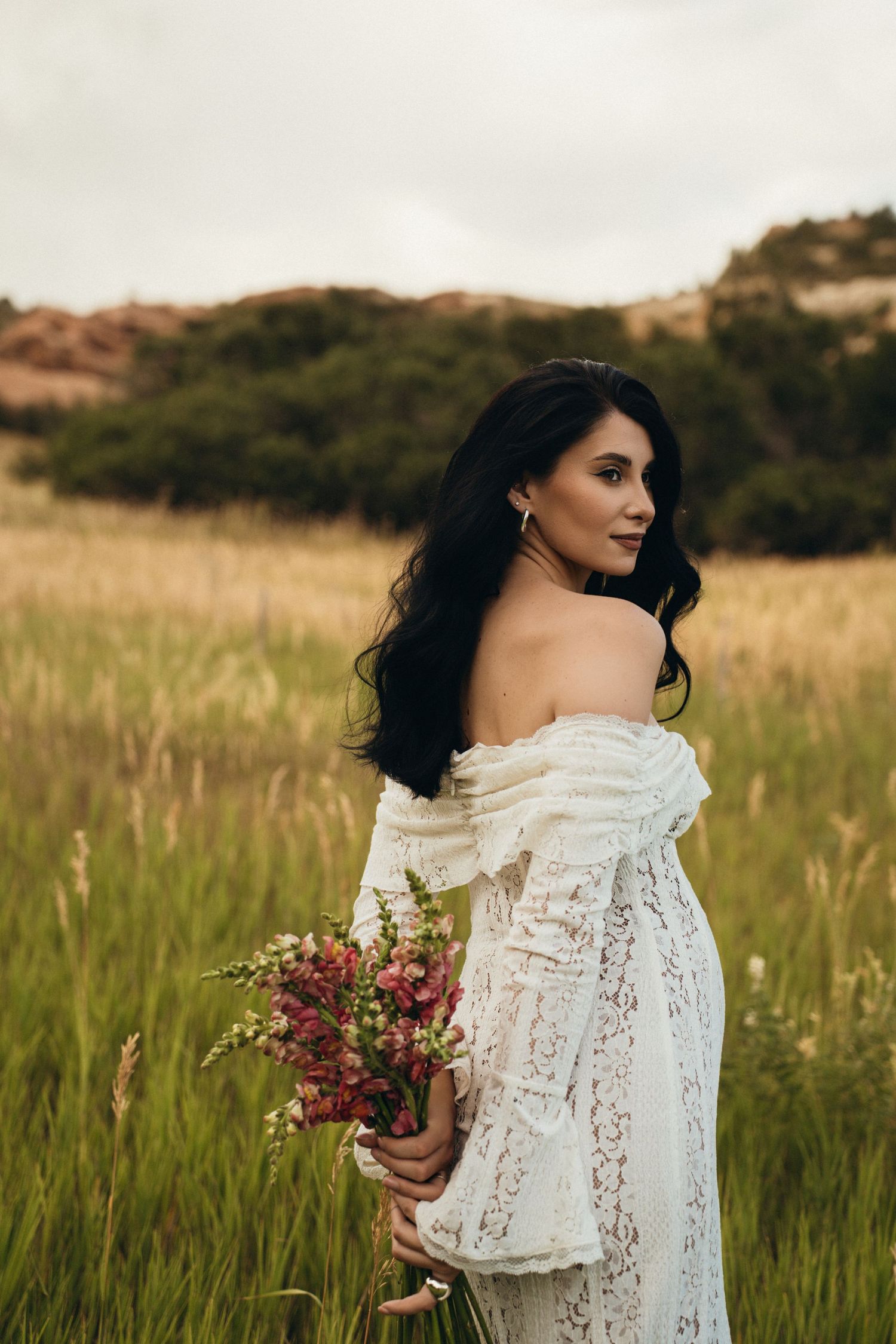 A person in a white lace off-shoulder dress holds a bouquet of red flowers while standing in a golden grassy field.