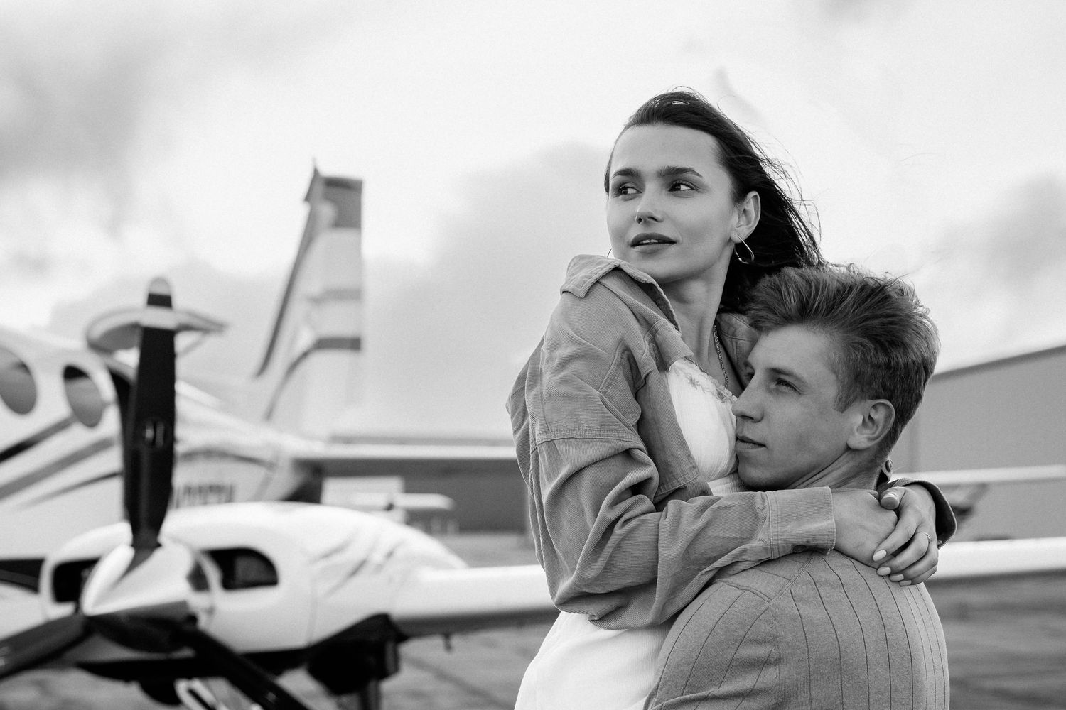 A romantic black and white photo showing a couple embracing near a small aircraft on an airfield.