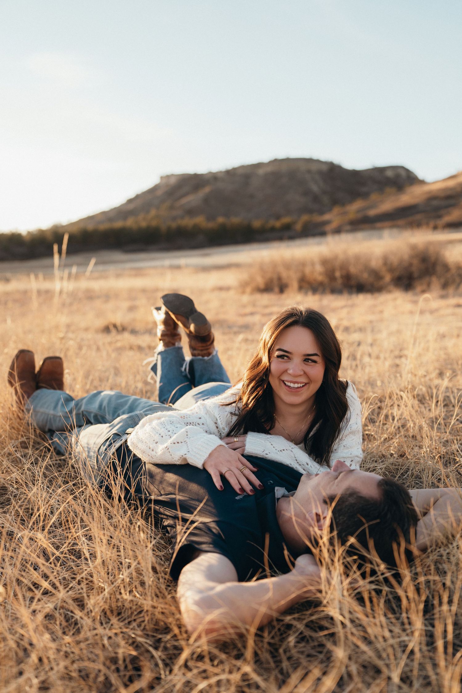 A couple relaxes together in golden grass with mountains in the background during sunset.