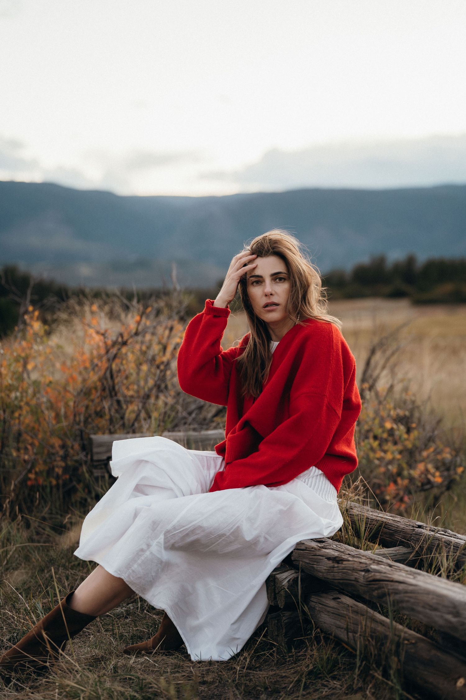 Person in red sweater and white dress poses gracefully on fallen log in scenic mountain landscape at sunset.