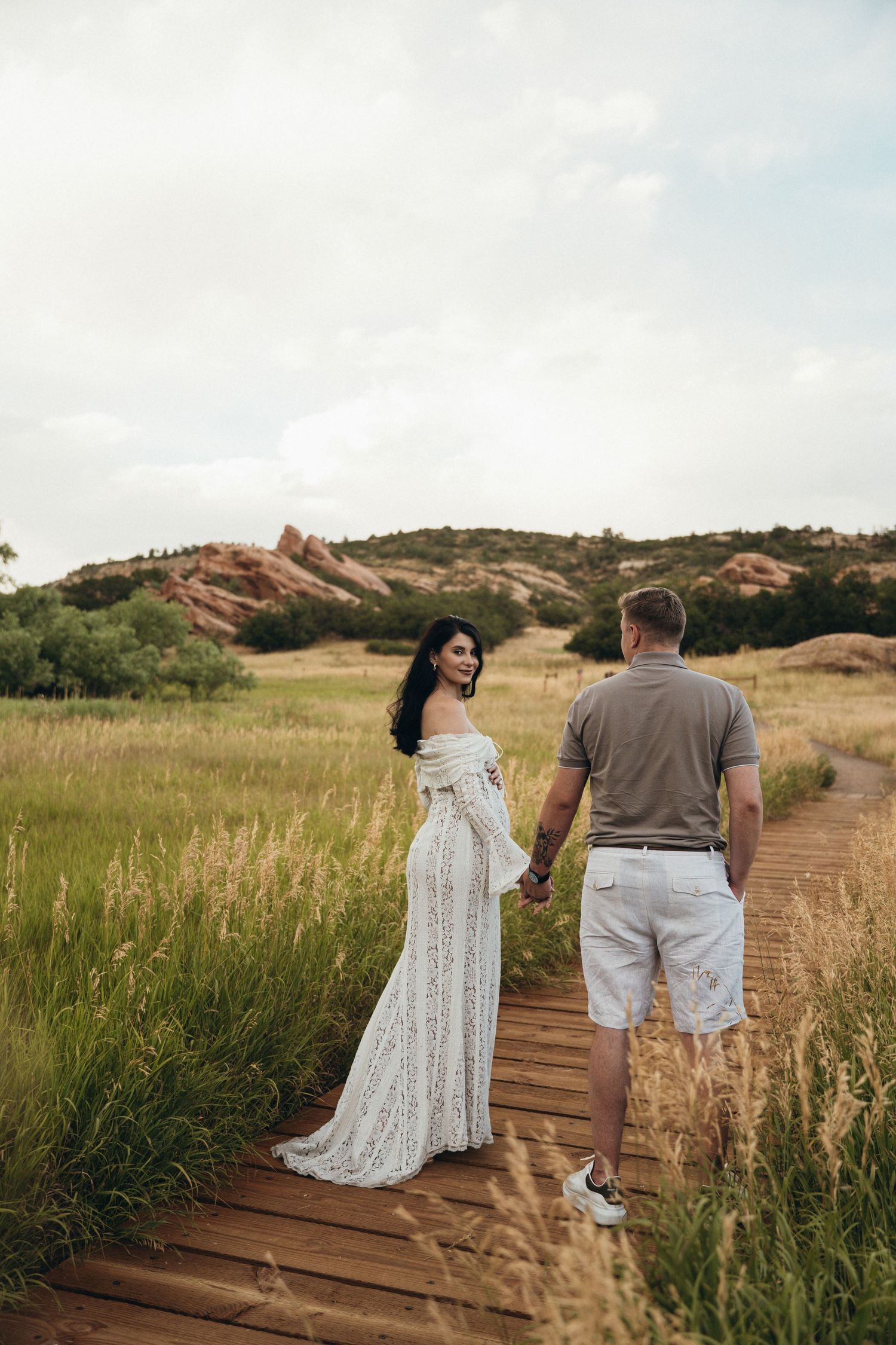 A couple stands on a wooden pathway through tall grass with red rock formations in the background at sunset.