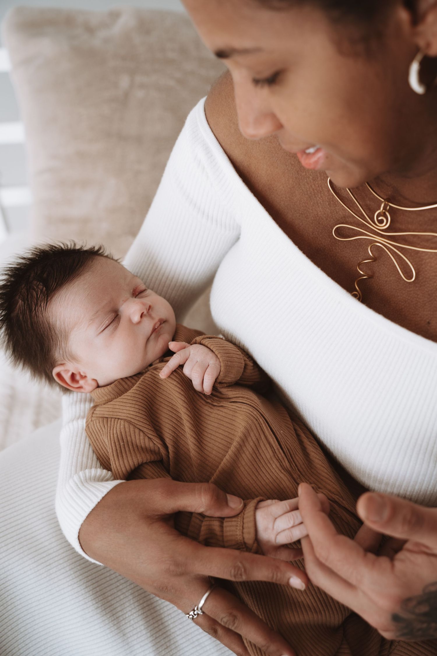 A sleeping newborn baby in a brown outfit rests peacefully in caring arms on soft white fabric.