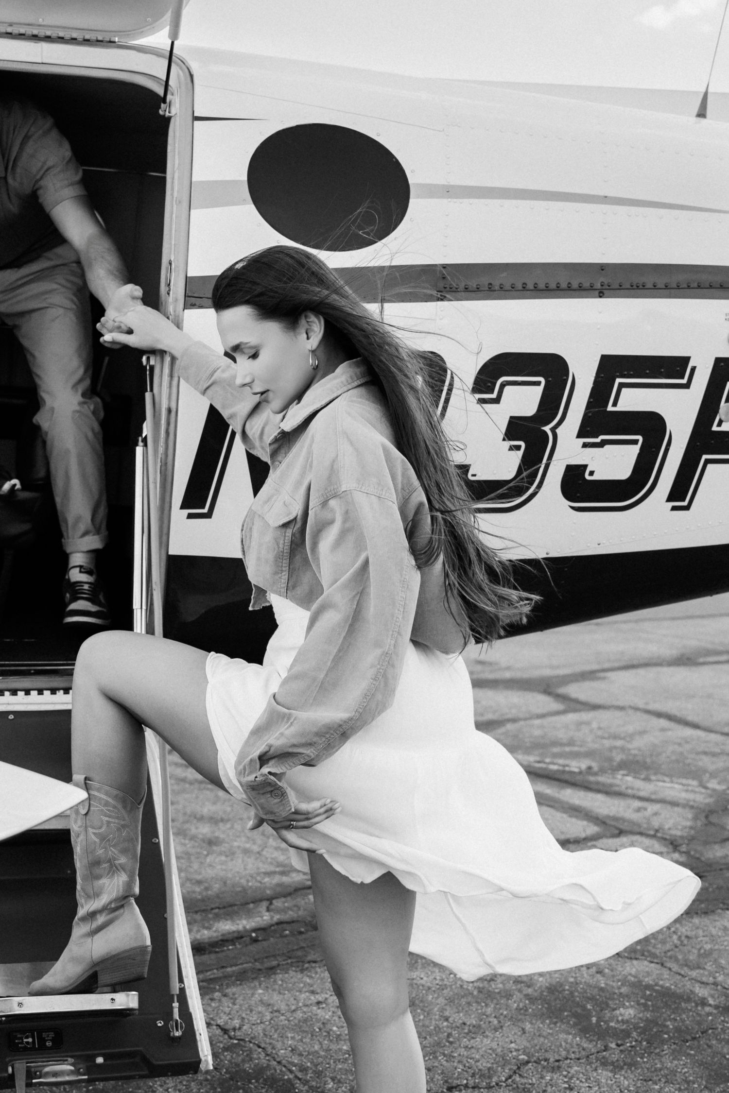 A woman in a light jacket boards an aircraft in a flowing white dress as her hair blows in the wind.