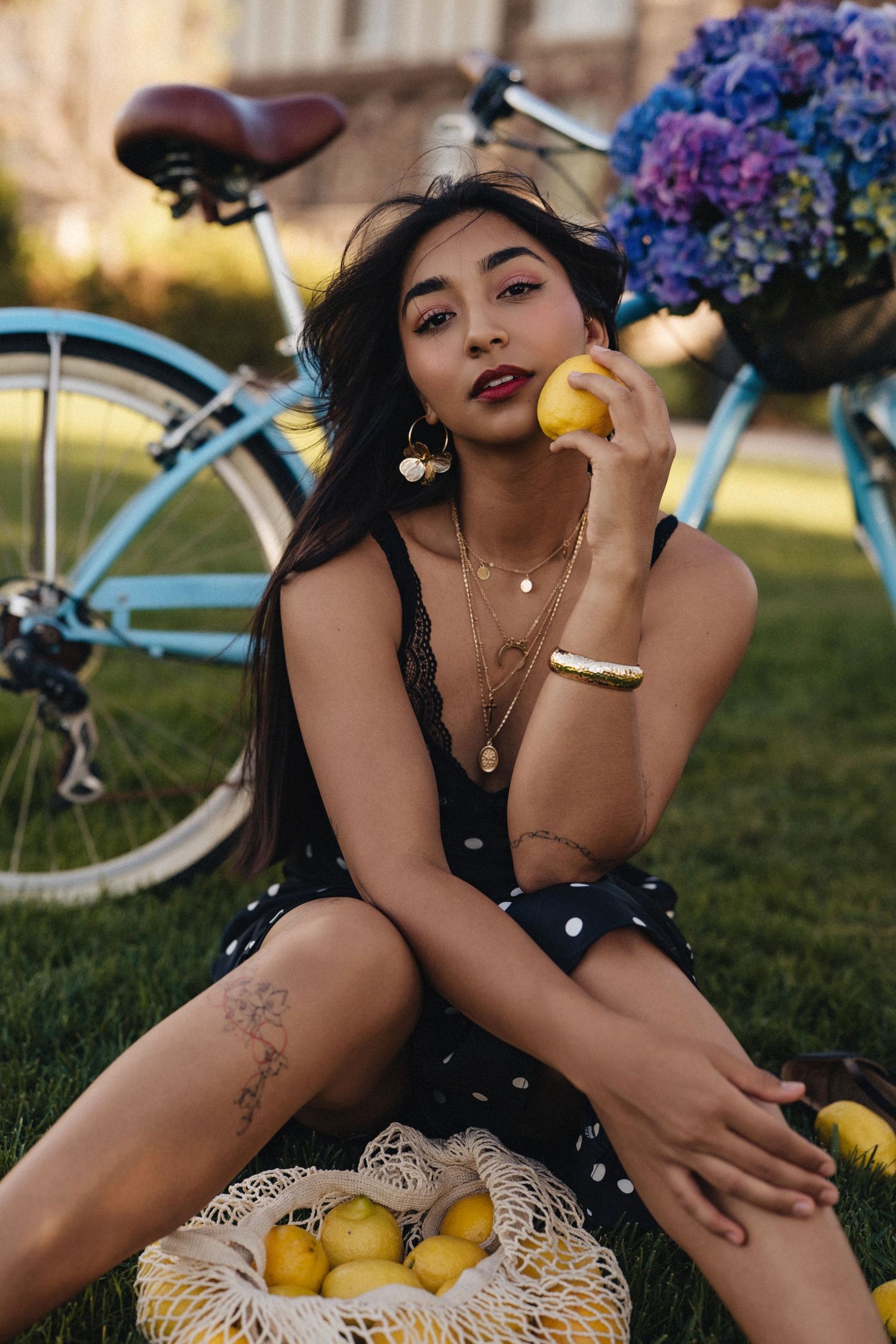 Young woman in polka dot dress sitting on grass holding a lemon, with a blue bicycle and purple flowers behind her.