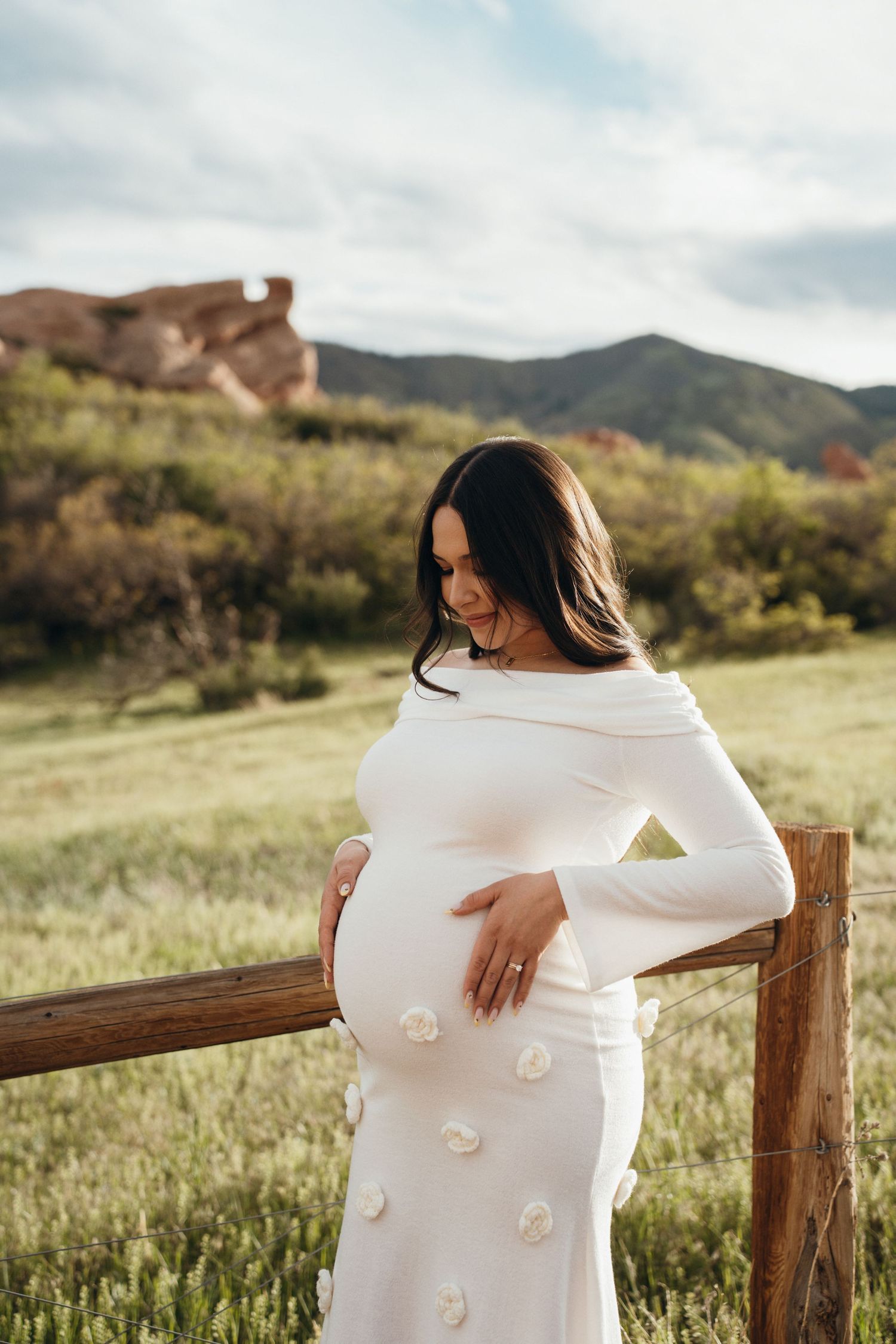 A pregnant woman in a white dress stands by a wooden fence in a scenic mountain landscape during golden hour.