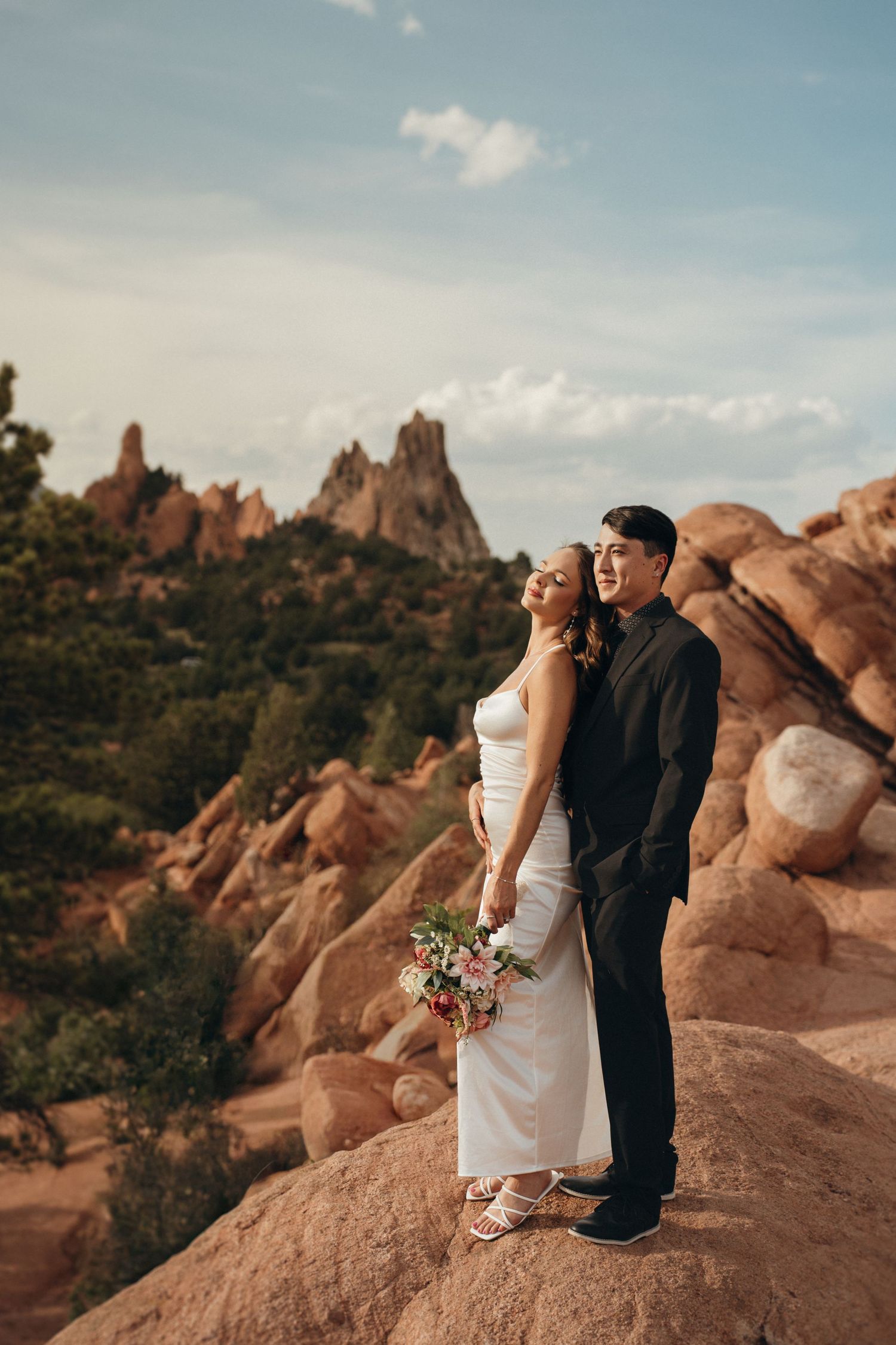 Wedding couple stands atop red rock formations at sunset in Garden of the Gods with dramatic mountain landscape views.