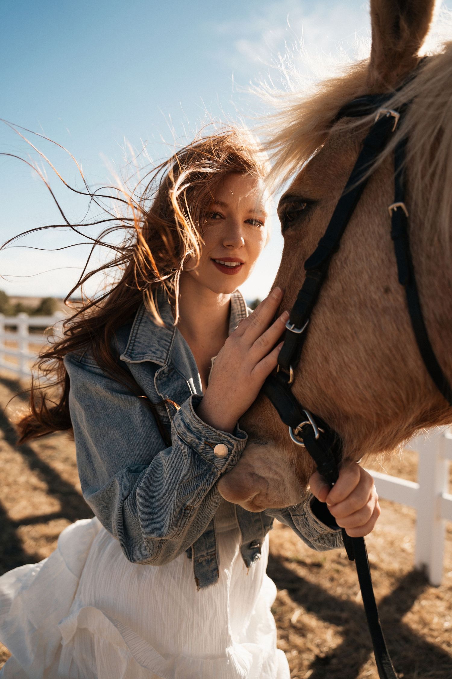 A woman in denim jacket and white skirt stands alongside a brown horse in warm sunlight.