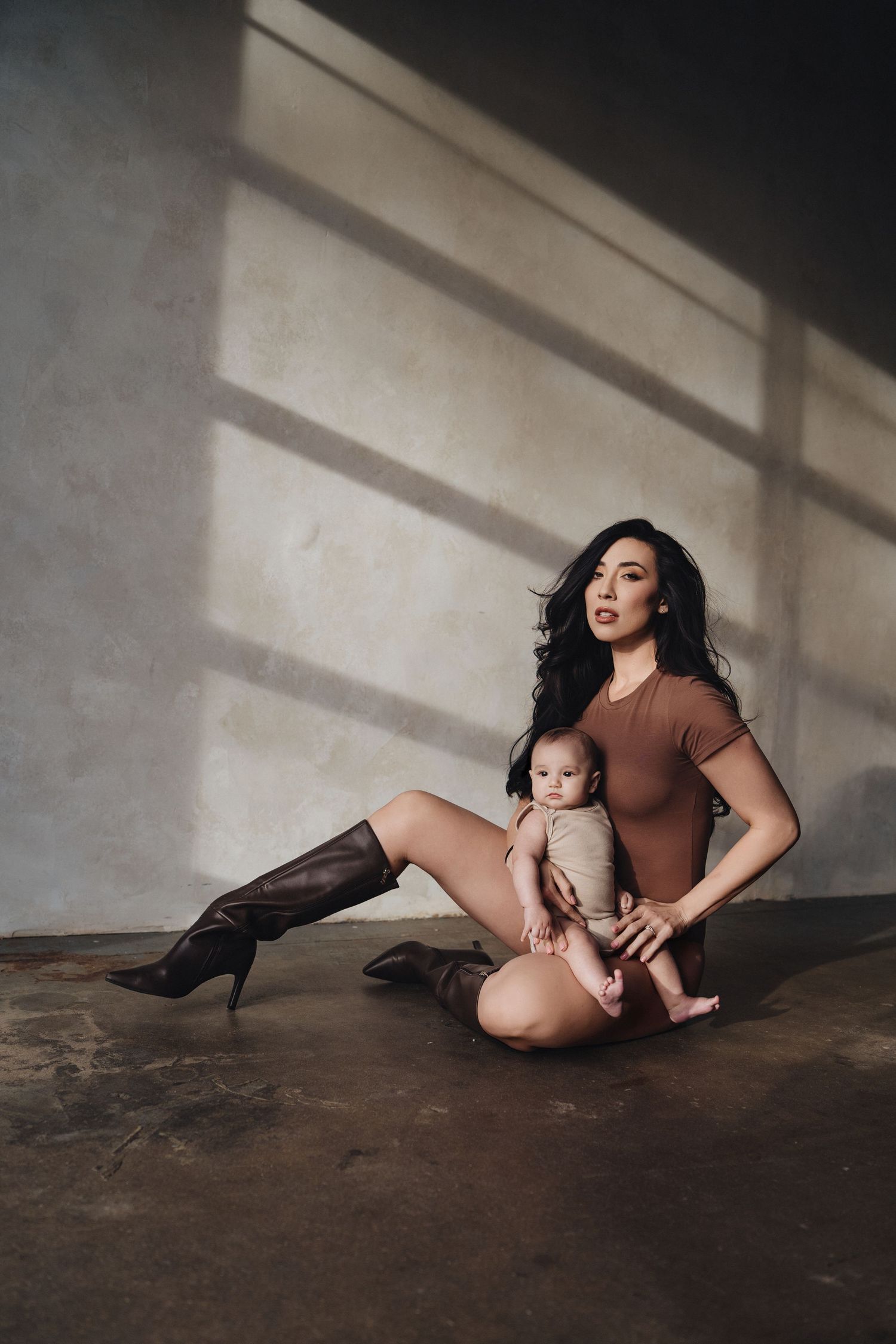 A parent and baby sit on a concrete floor in dramatic lighting with shadows from a window falling on the wall.