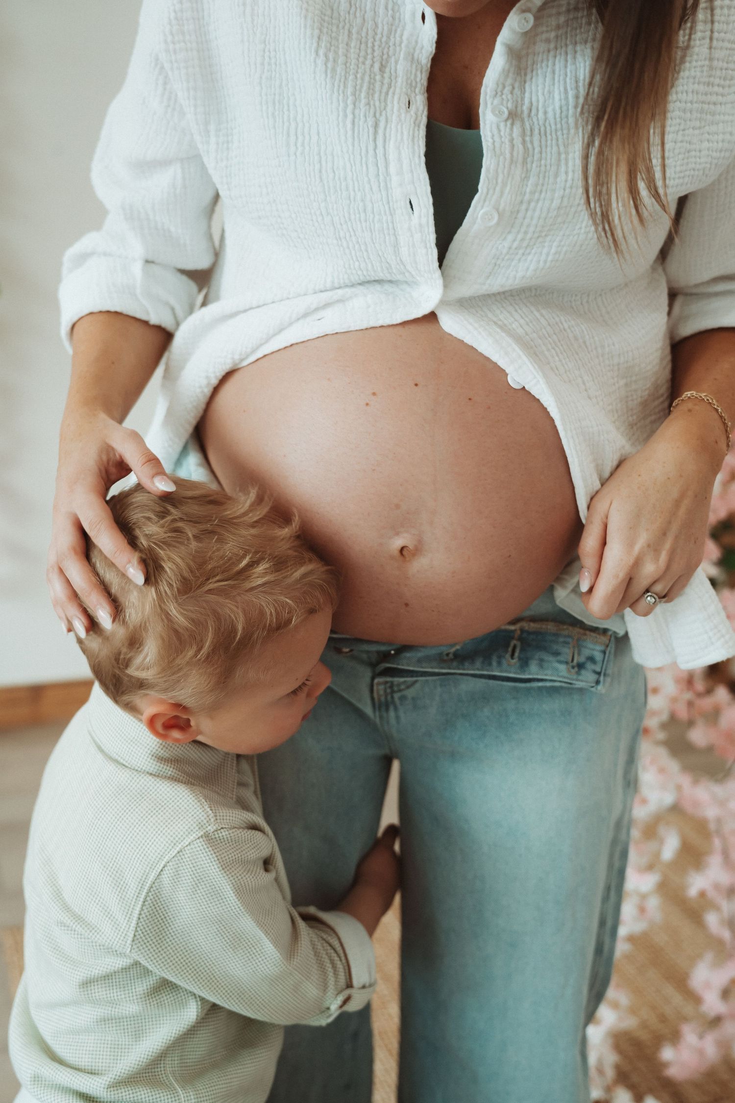 A toddler in white sweater kisses pregnant belly while mother wears unbuttoned white shirt and jeans.