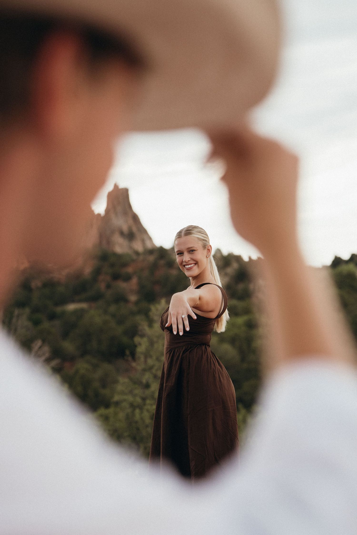 Smiling blonde woman in dark dress showing engagement ring outdoors with rocky mountain landscape background.