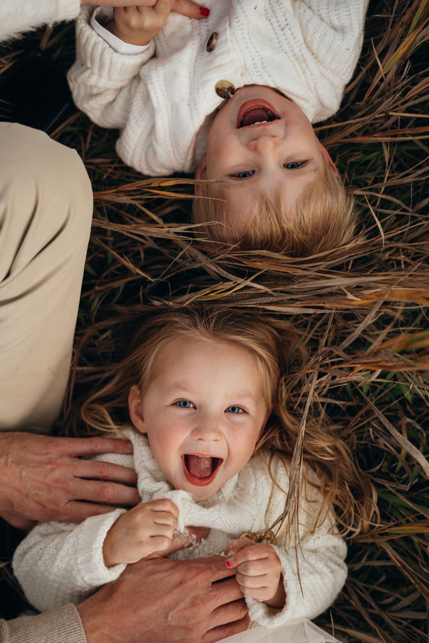 A joyful child lies in dry grass wearing a white sweater while someone's hands gently touch their face.