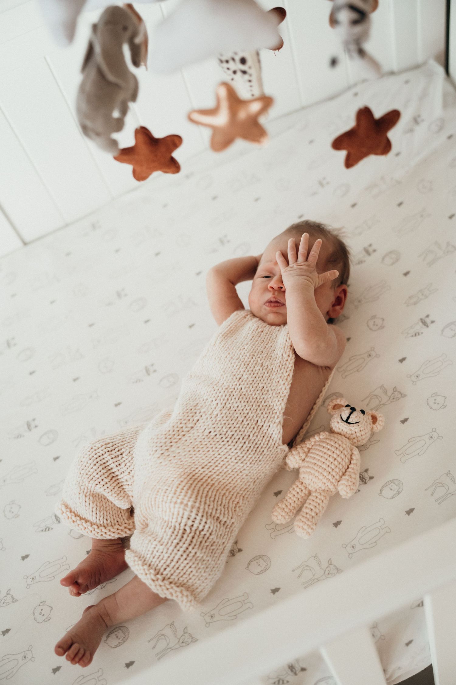 Sleeping baby in cream knit romper lying on patterned bedding with stuffed bear toy and hanging star decorations above.