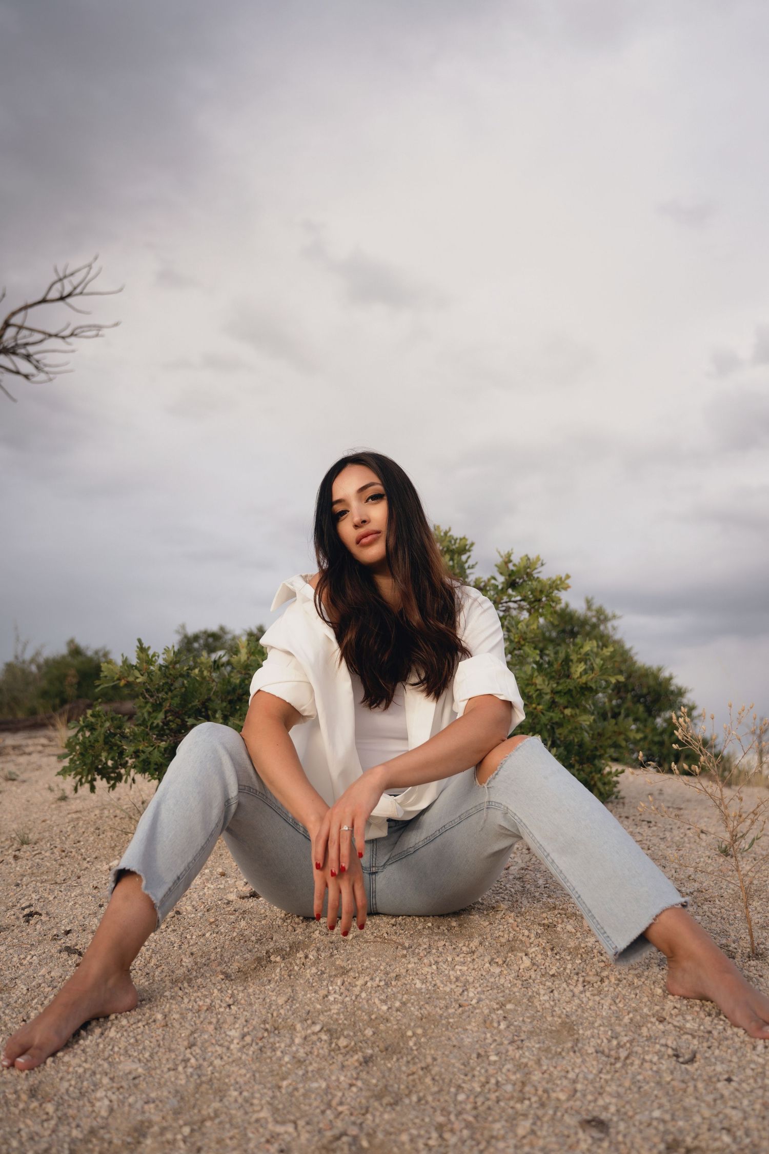Woman sits barefoot on rocky ground against dramatic cloudy sky.