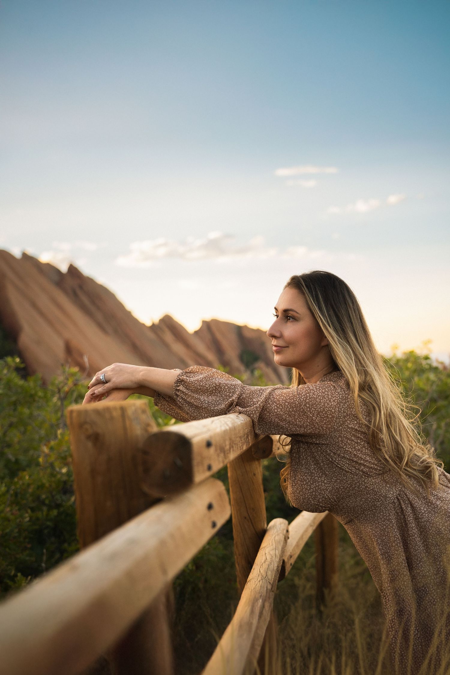 Woman in brown dress leaning on wooden railing at sunset with mountains.