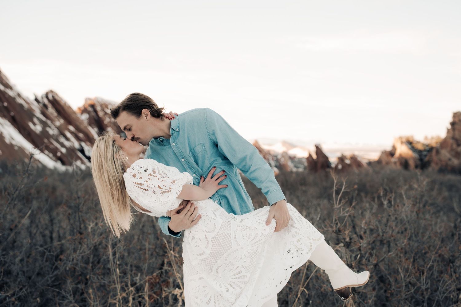 A romantic couple in a white dress and turquoise shirt embrace in a dip pose against a dusky natural landscape.