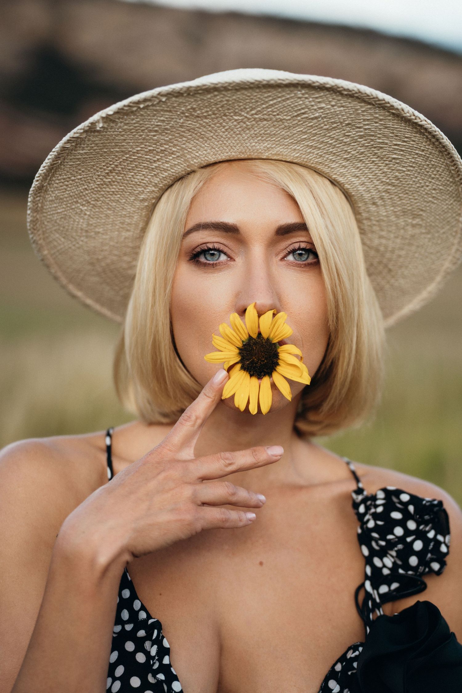 Blonde woman in straw hat holding a yellow sunflower near her face, wearing a black polka dot dress outdoors.