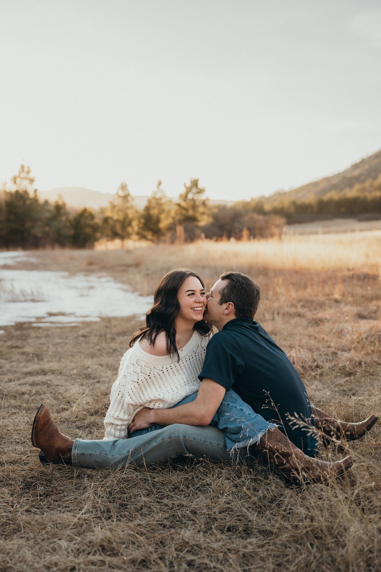 A couple lies together in a golden field at sunset sharing an intimate moment near mountains and water.