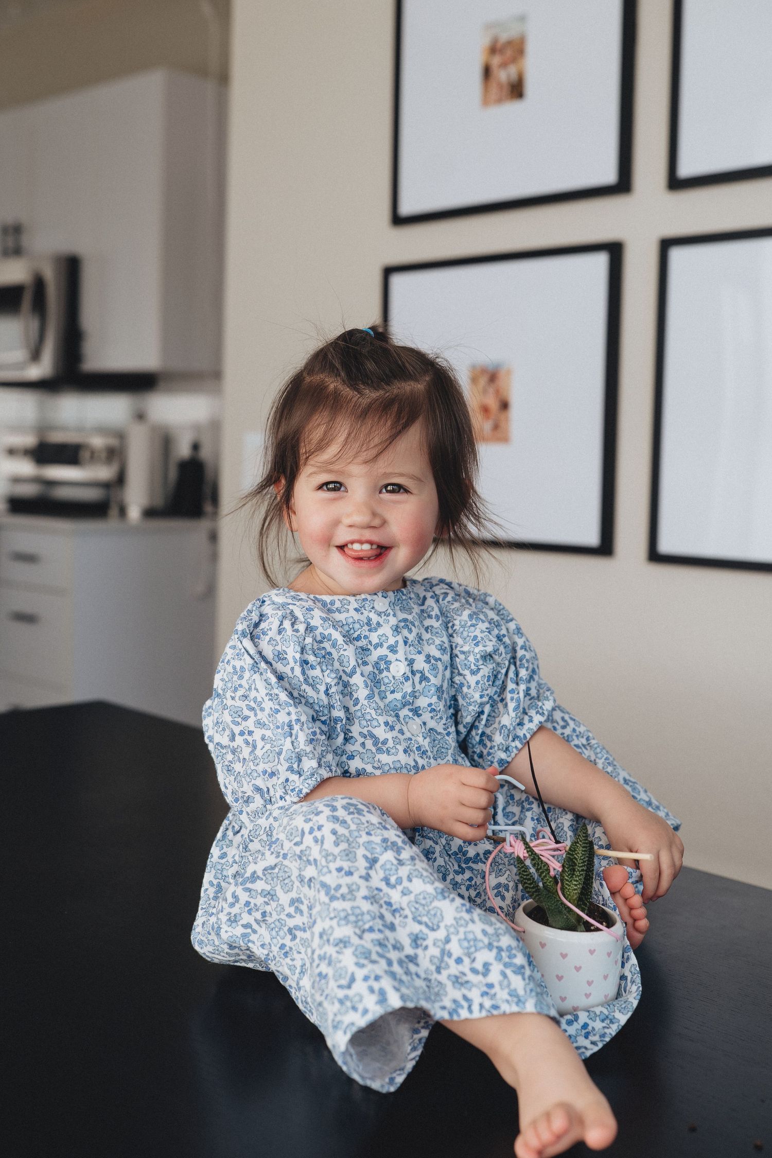 Smiling toddler girl in blue floral dress sitting on table holding small potted plant in modern home.