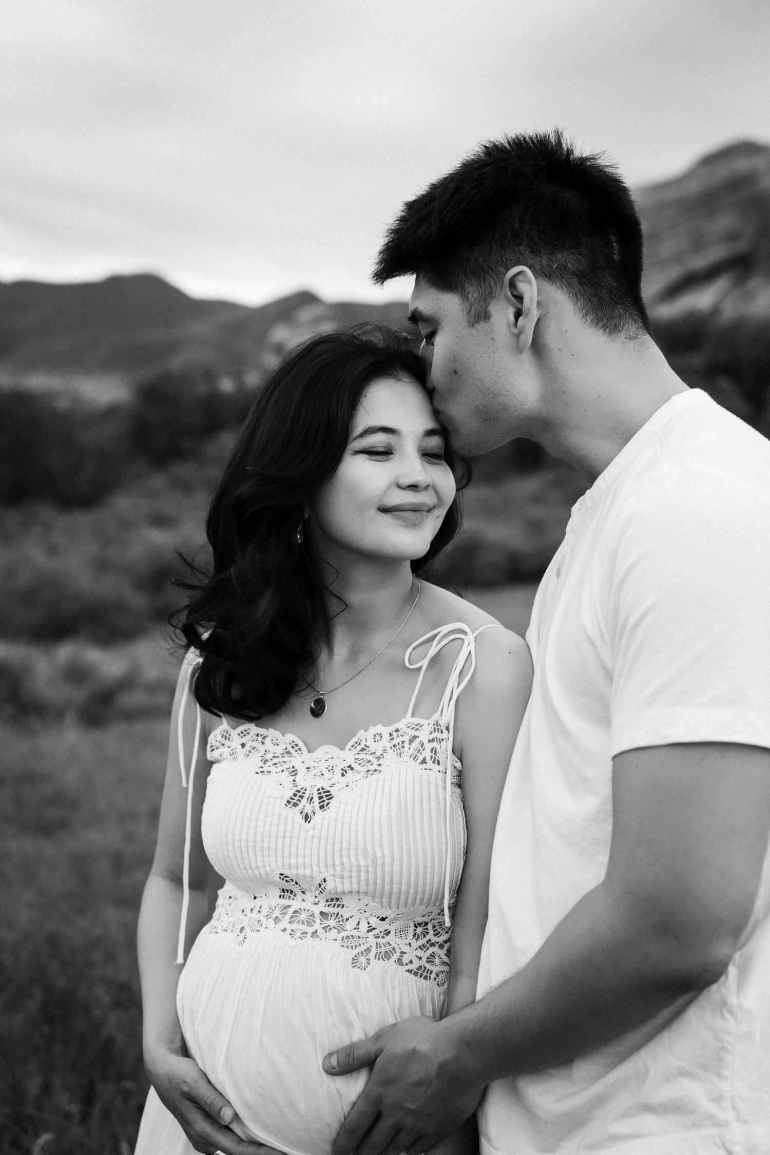 Black and white photo of a man kissing a pregnant woman's forehead outdoors with mountains in the background.