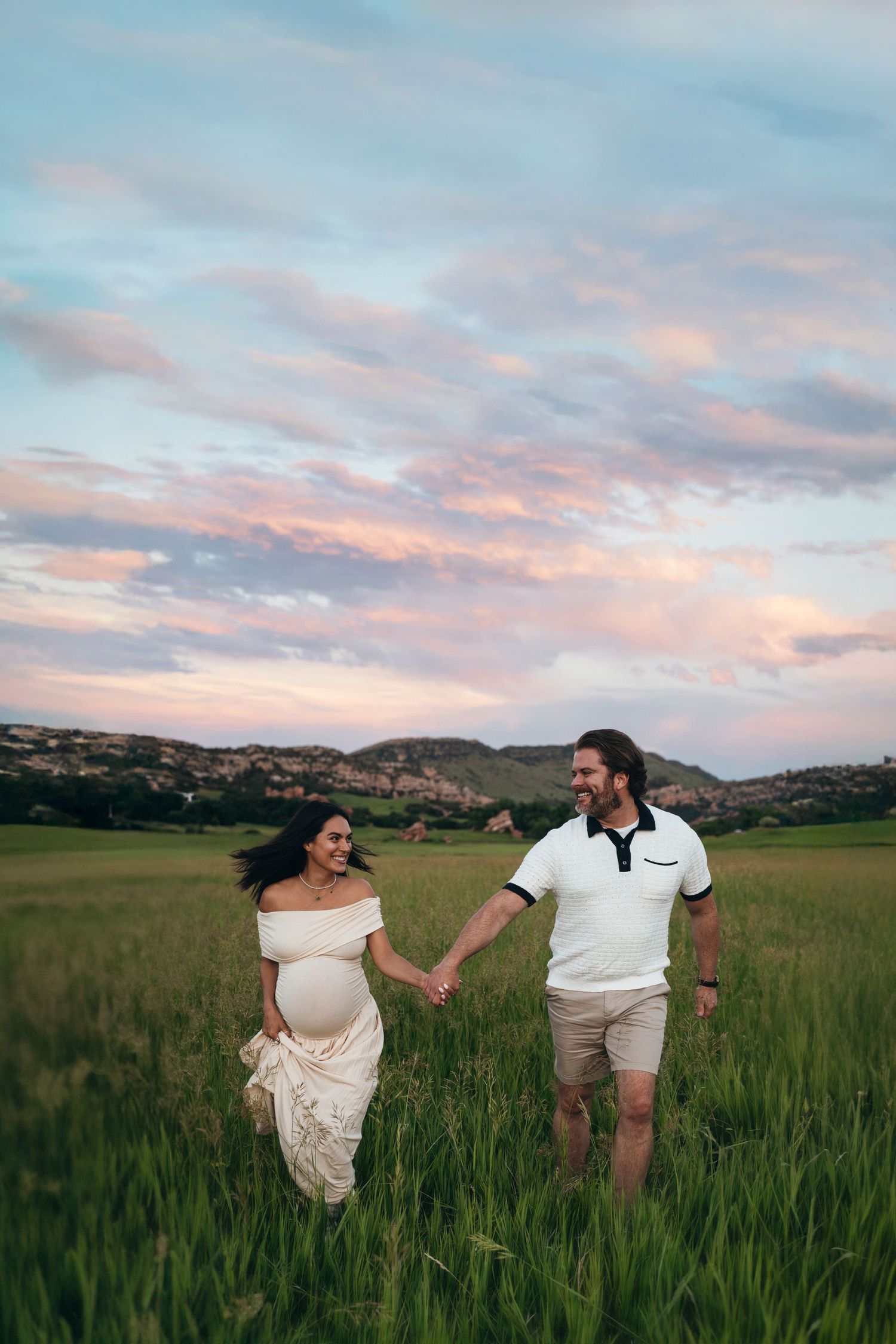 Two people hold hands while walking through a green meadow at sunset with mountains in the background.